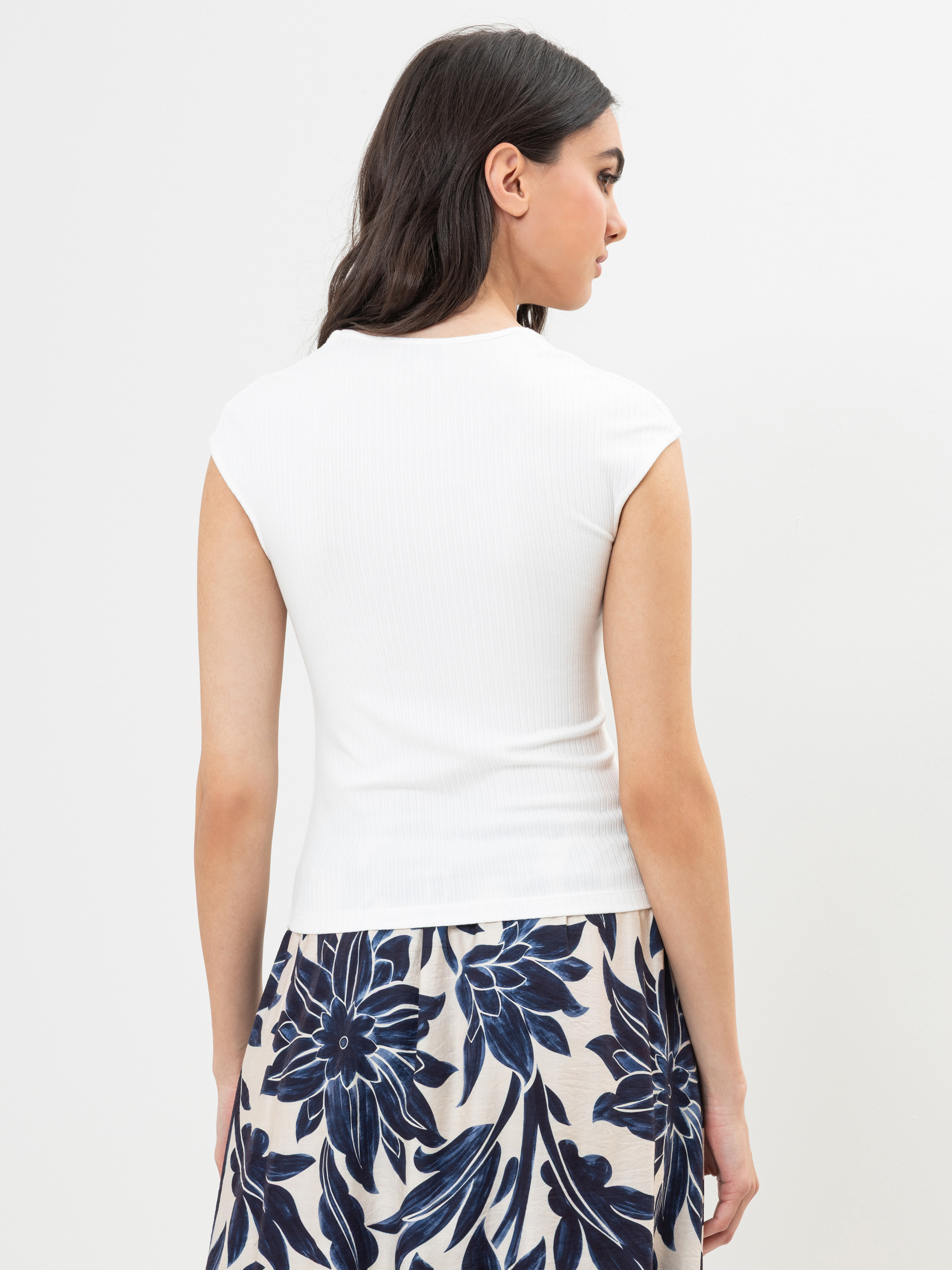 Eine Frau mit langen dunklen Haaren blickt von sich weg, trägt ein weißes Oberteil mit langen Ärmeln und einen cremefarbenen Rock mit einem großen blauen Blumenmuster und steht vor einem schlichten hellen Hintergrund.