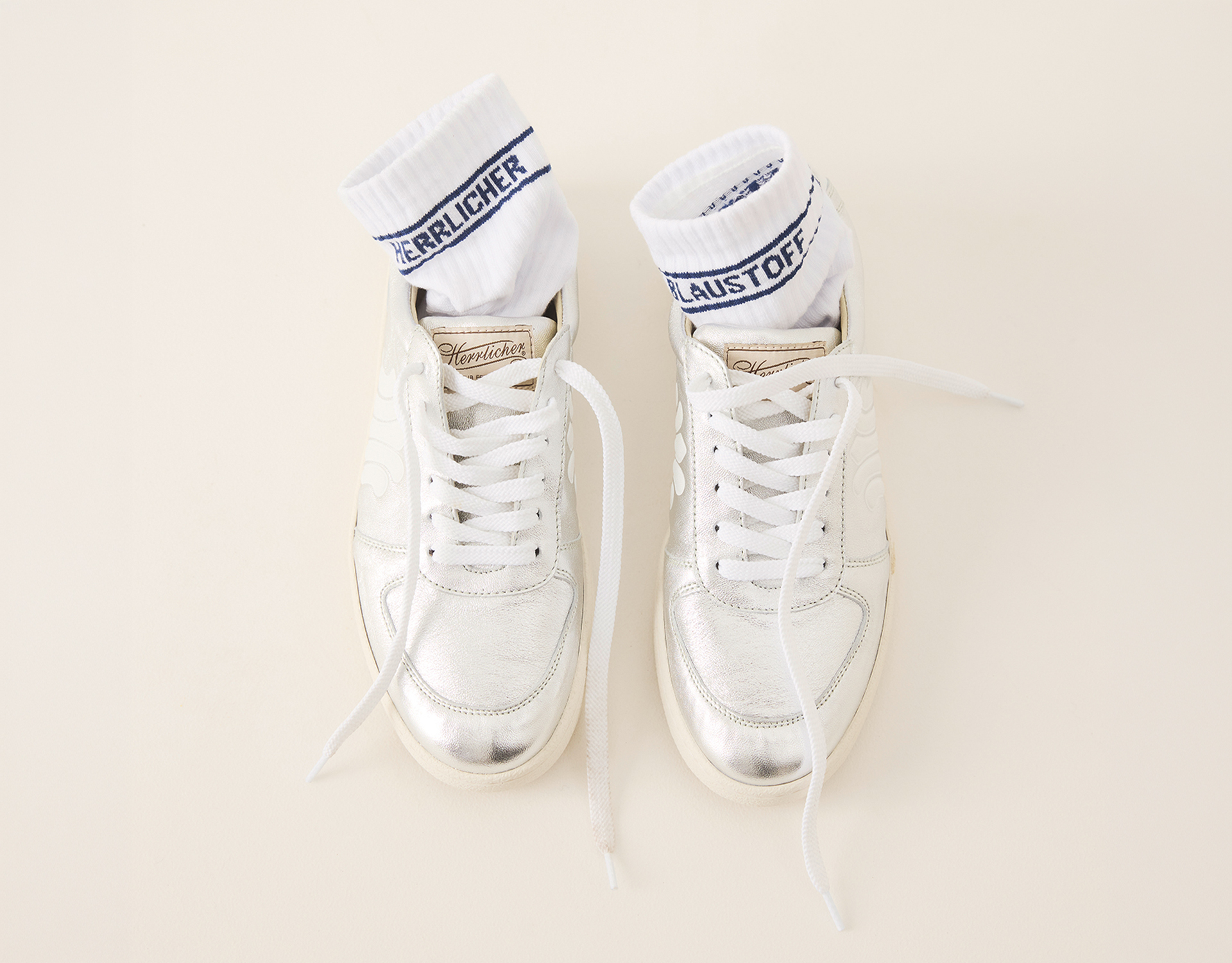 Ein Paar weißer Turnschuhe mit silbernen Metallic-Akzenten, wobei jeder Schuh eine aufgerollte weiße Socke mit blauem Text auf dem Bündchen enthält, die auf einer hellbeigen Fläche steht.