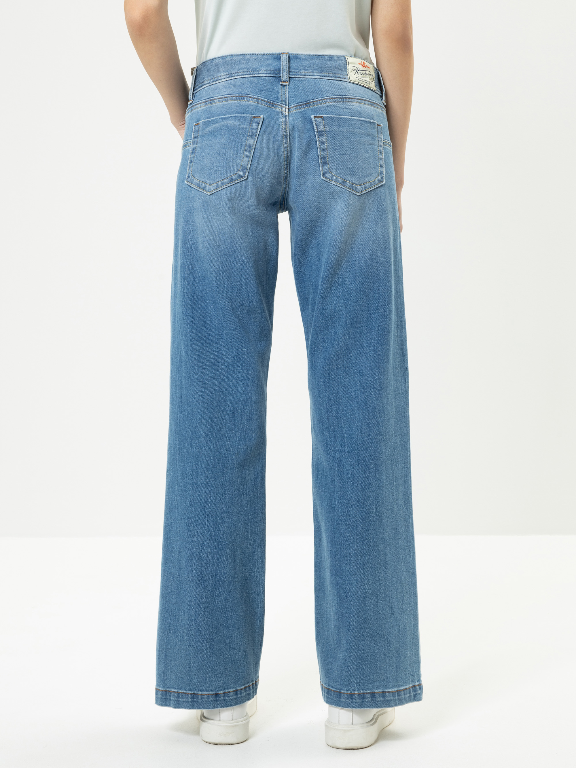 Eine Person steht vor einem schlichten Hintergrund und trägt eine weit geschnittene blaue Jeans und weiße Schuhe. Das Foto ist von hinten aufgenommen und konzentriert sich auf die Gesäßtaschen und die lockere Passform der Jeans.