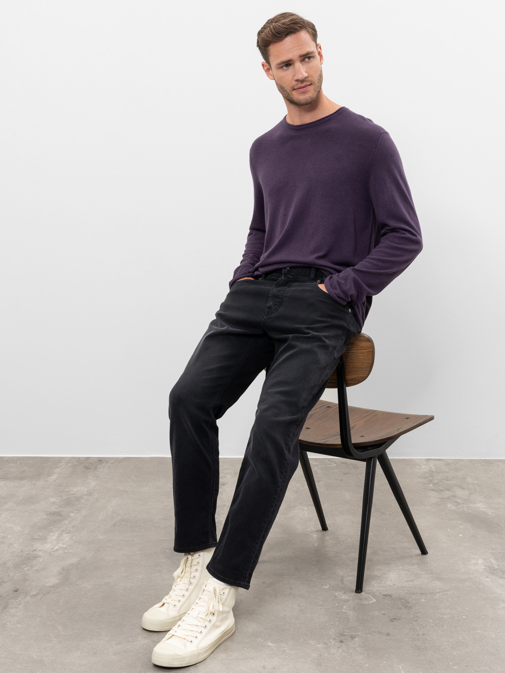 Ein Mann in einem lilafarbenen langärmeligen Hemd, einer schwarzen Hose und weißen Turnschuhen sitzt seitwärts auf einem Holzstuhl, die Hände in den Taschen, vor einer schlichten weißen Wand und einem grauen Betonboden.