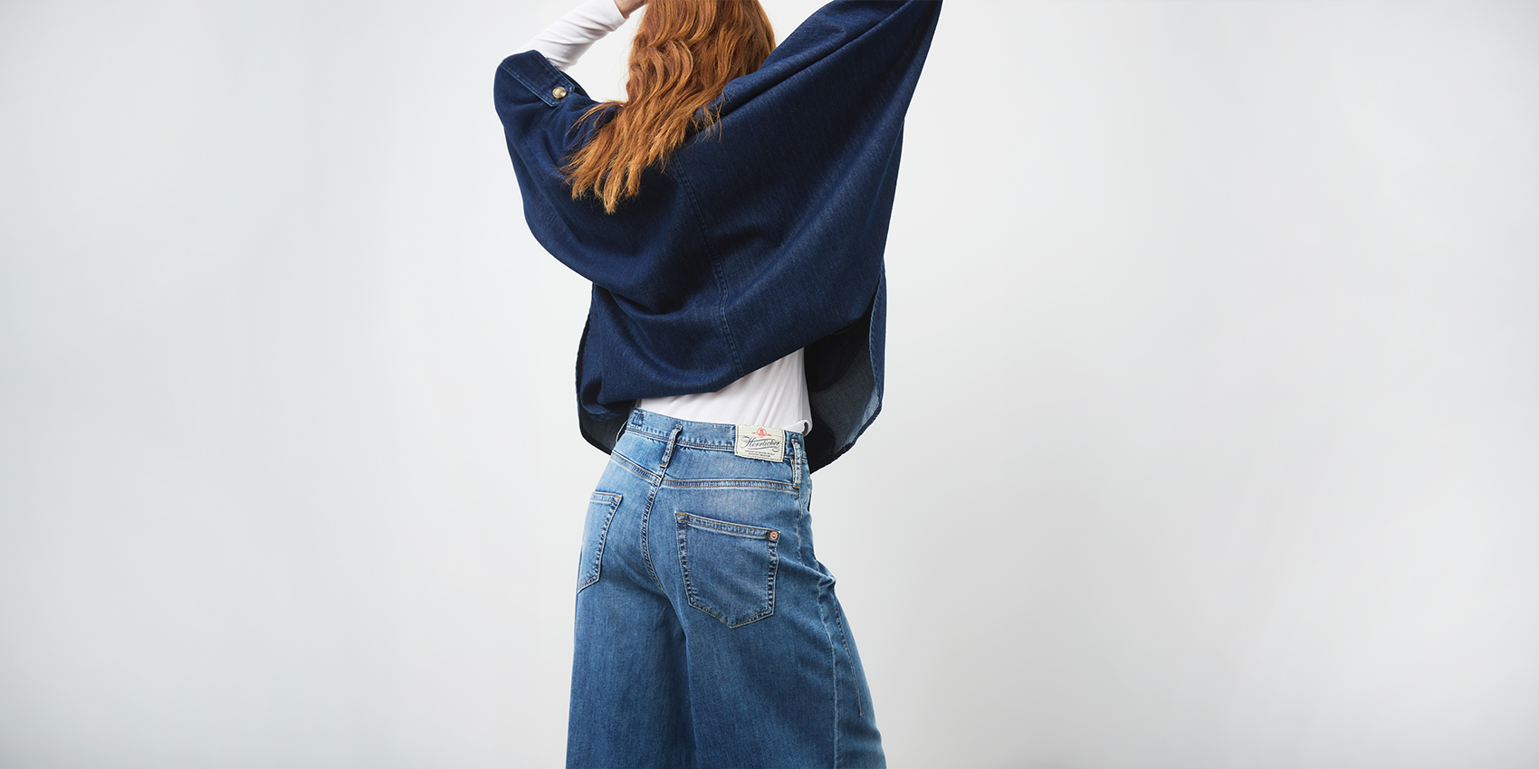 Eine Person mit langen roten Haaren, die einen dunkelblauen Poncho, ein weißes Oberteil und eine blaue Jeans mit weitem Bein trägt, steht mit dem Rücken zur Kamera und leicht erhobenen Armen vor einem schlichten, hellen Hintergrund.