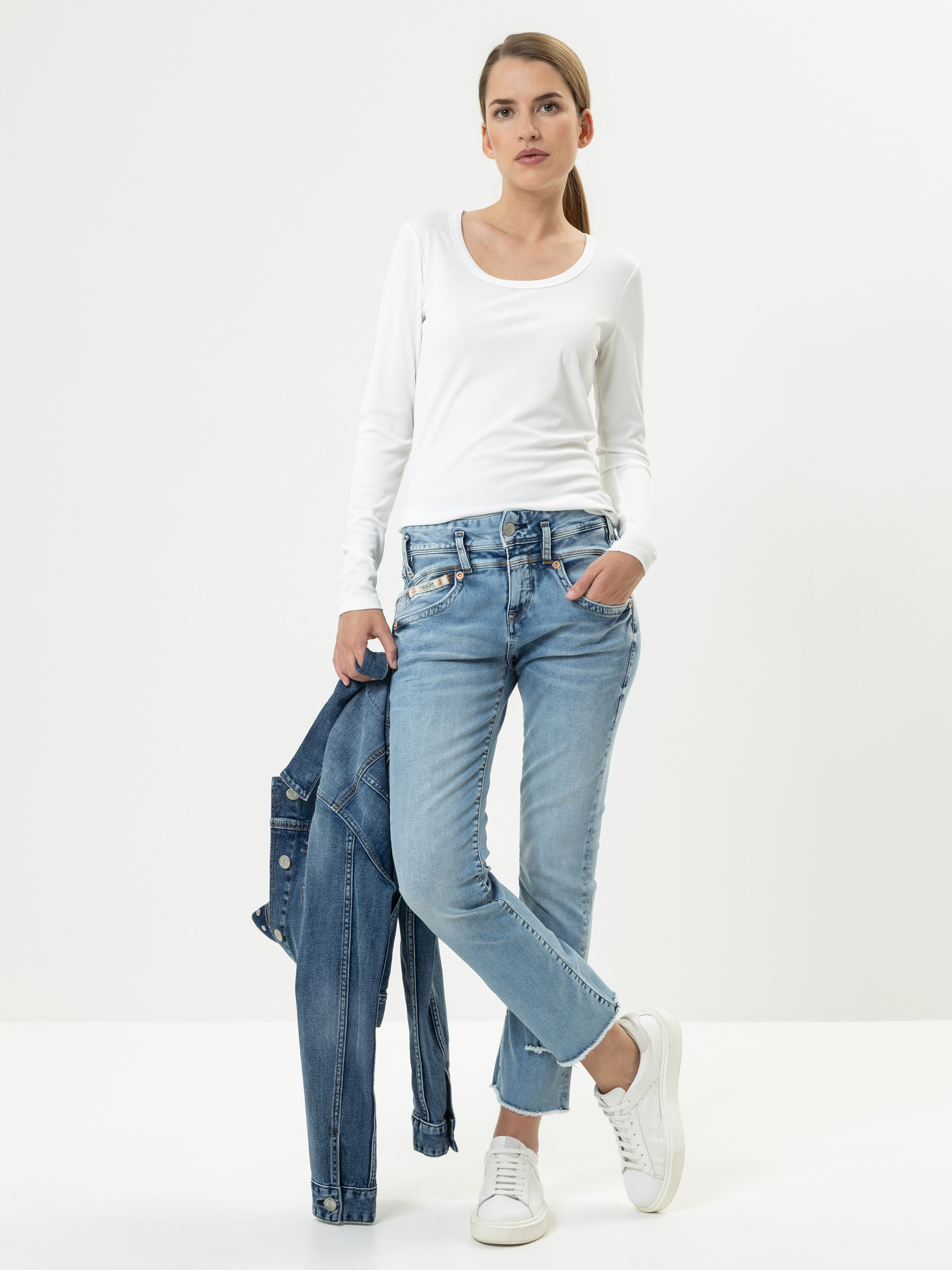 Eine Frau steht vor einem schlichten weißen Hintergrund und trägt ein weißes langärmeliges Oberteil, hellblaue Jeans und weiße Turnschuhe. Sie hält eine Jeansjacke in der einen Hand und hat die andere Hand in der Tasche.