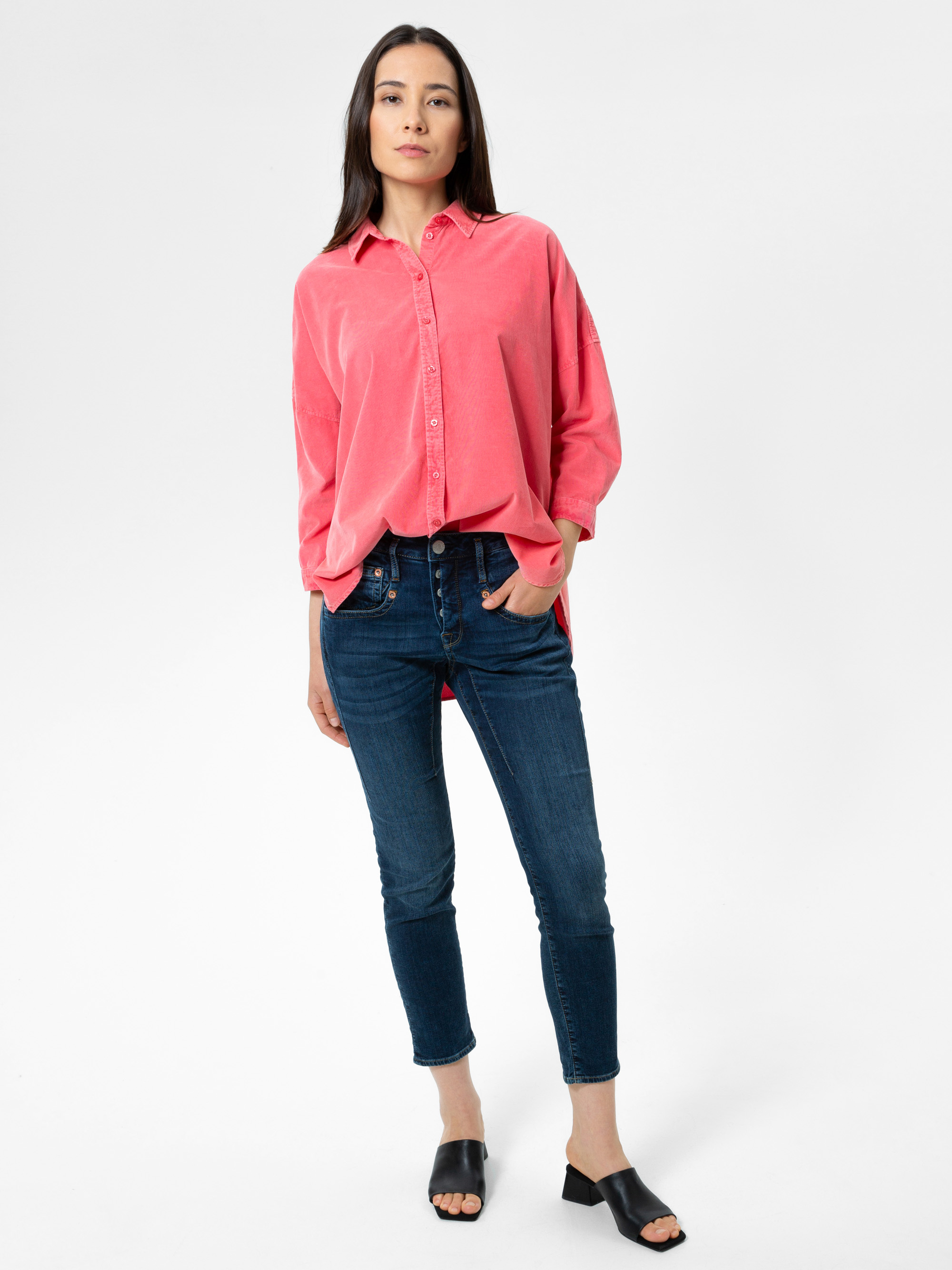 Eine Frau steht vor einem schlichten weißen Hintergrund. Sie trägt ein lockeres rosafarbenes Button-up-Hemd, dunkelblaue Skinny-Jeans und schwarze Sandalen mit offenen Zehenabsätzen. Sie hat lange dunkle Haare und eine Hand in der Tasche.