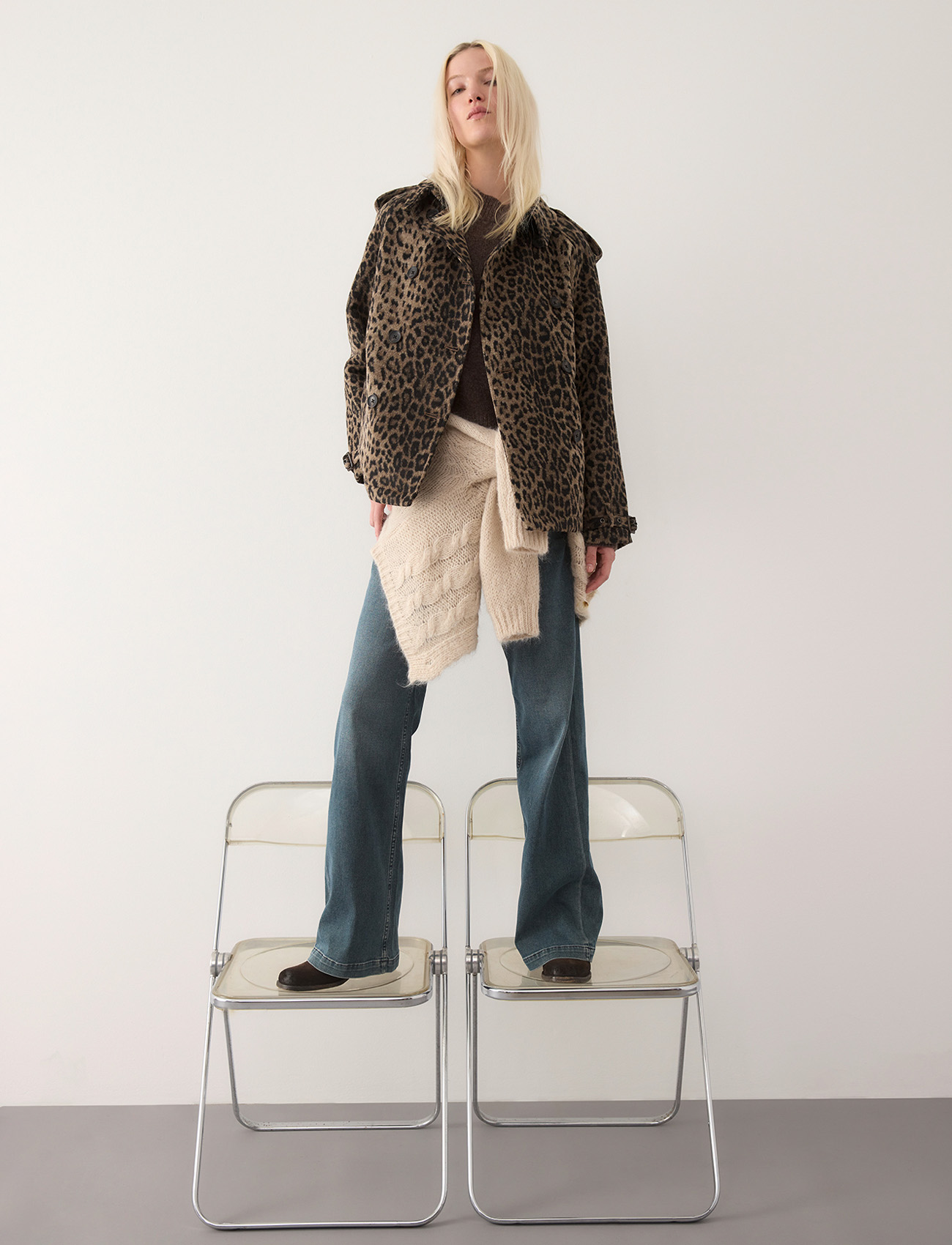 Eine Person mit langen blonden Haaren steht auf zwei durchsichtigen Klappstühlen, trägt eine Jacke mit Leopardenmuster, blaue Jeans, schwarze Schuhe und einen beigen Pullover, der um die Taille gebunden ist, vor einer schlichten weißen Wand.