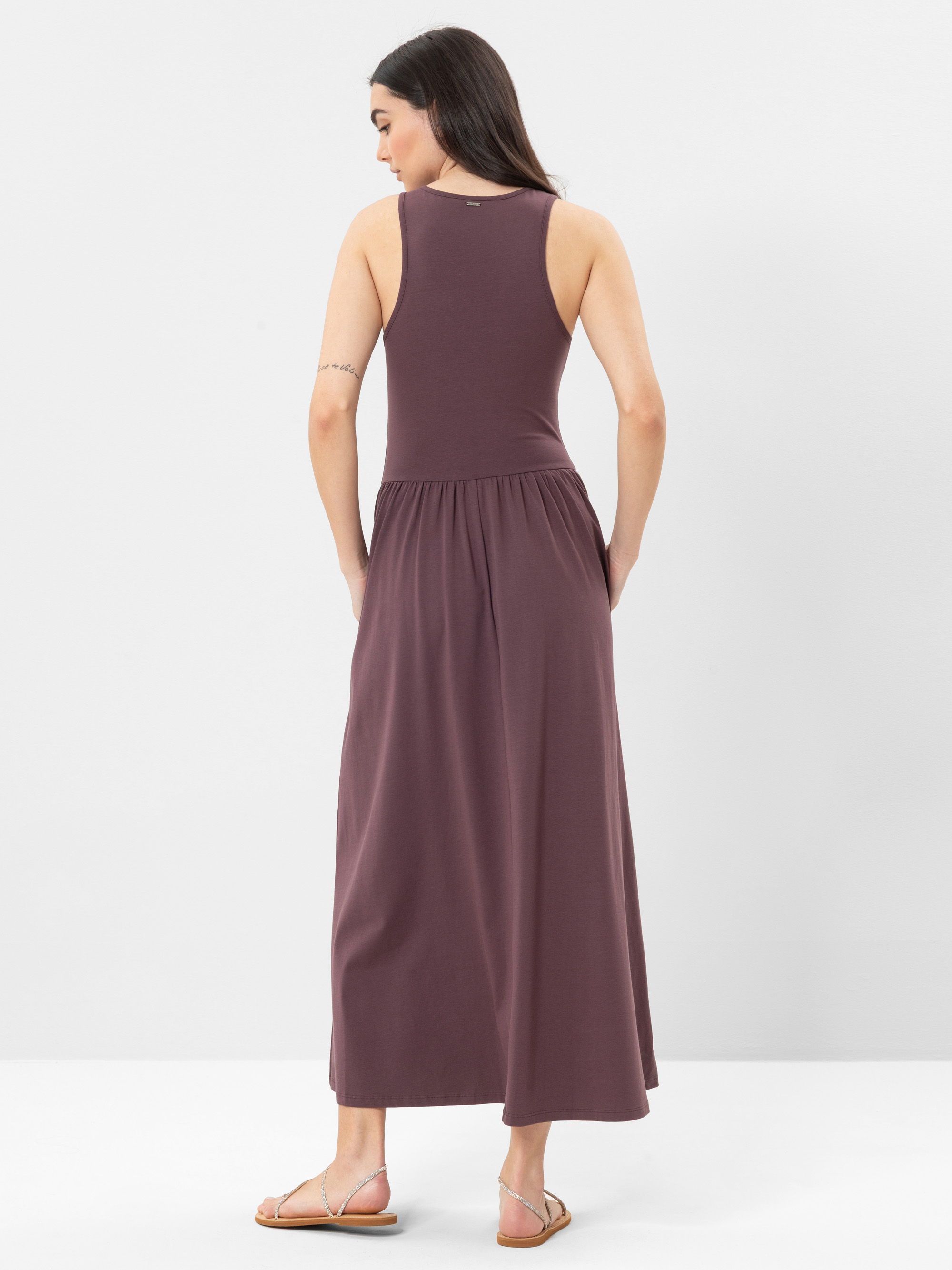 Eine Frau mit langen braunen Haaren, die ein ärmelloses, knöchellanges lila Kleid und Sandalen trägt, steht mit dem Rücken zur Kamera vor einem schlichten weißen Hintergrund.