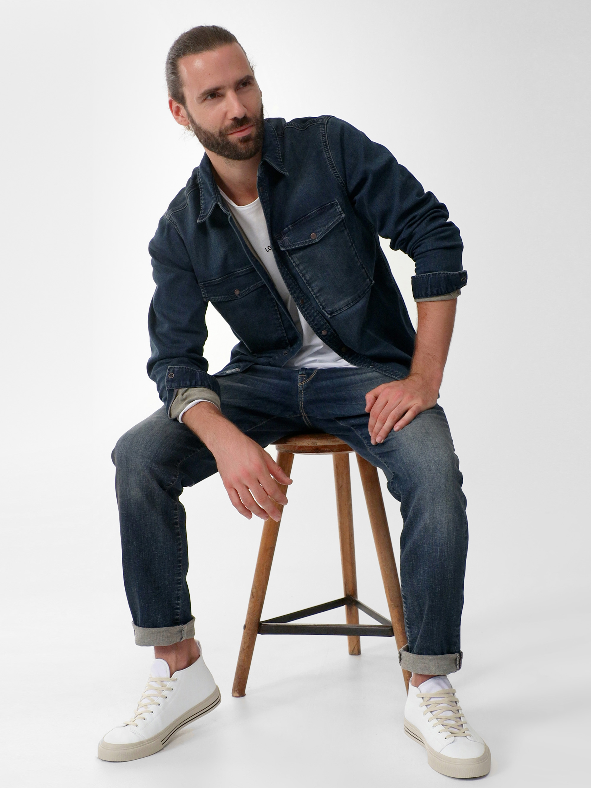 Ein bärtiger Mann mit zurückgebundenen langen Haaren sitzt auf einem Holzschemel. Er trägt eine dunkle Jeansjacke, hochgekrempelte Jeans, ein weißes T-Shirt und weiße Turnschuhe und posiert vor einem schlichten weißen Hintergrund.