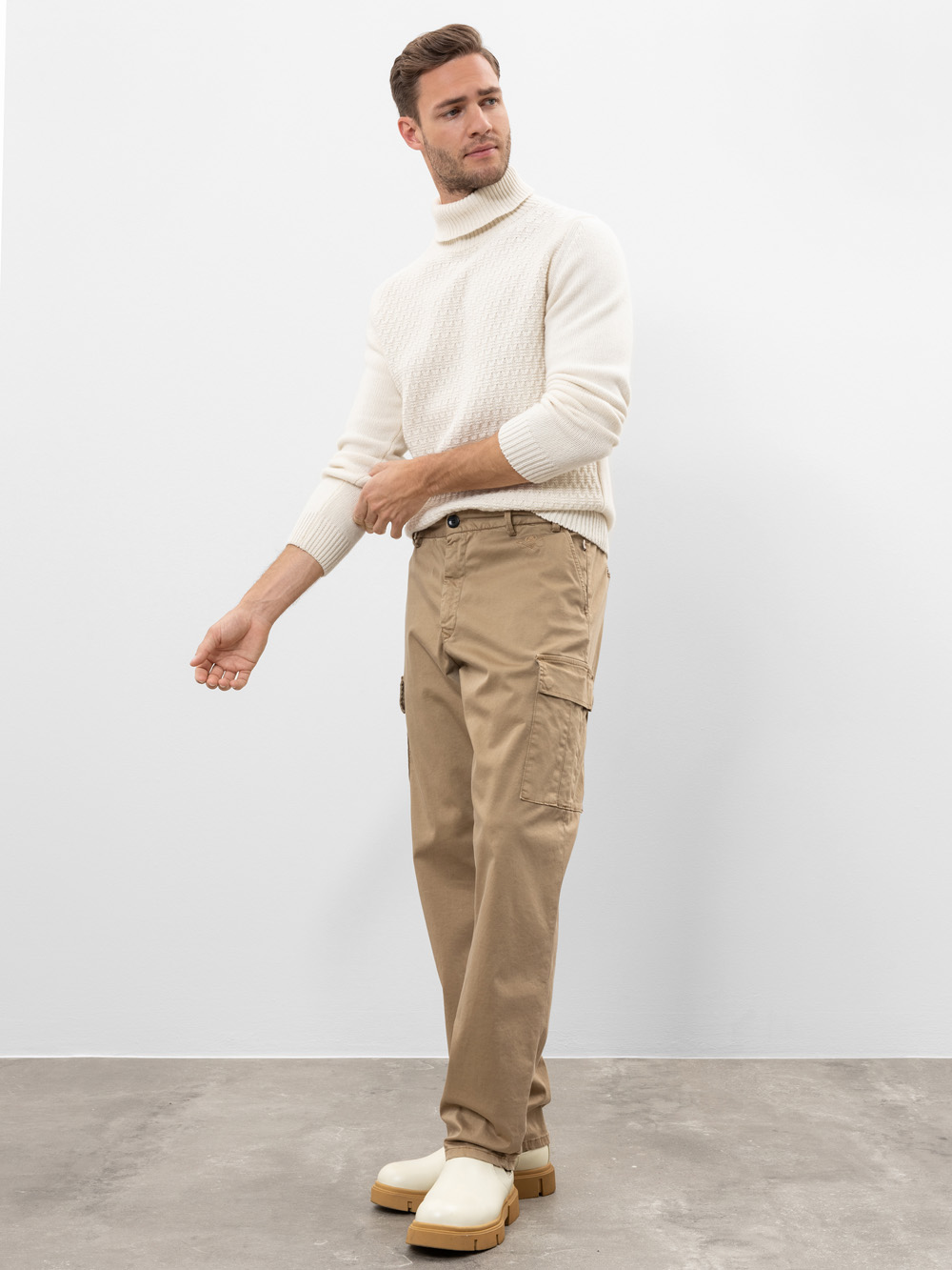Ein Mann steht auf einem grauen Fußboden vor einer schlichten weißen Wand. Er trägt einen cremefarbenen Rollkragenpullover, eine hellbraune Cargohose und wollweiße Schuhe. Er blickt mit ausgestrecktem Arm und entspannter Körperhaltung zur Seite.