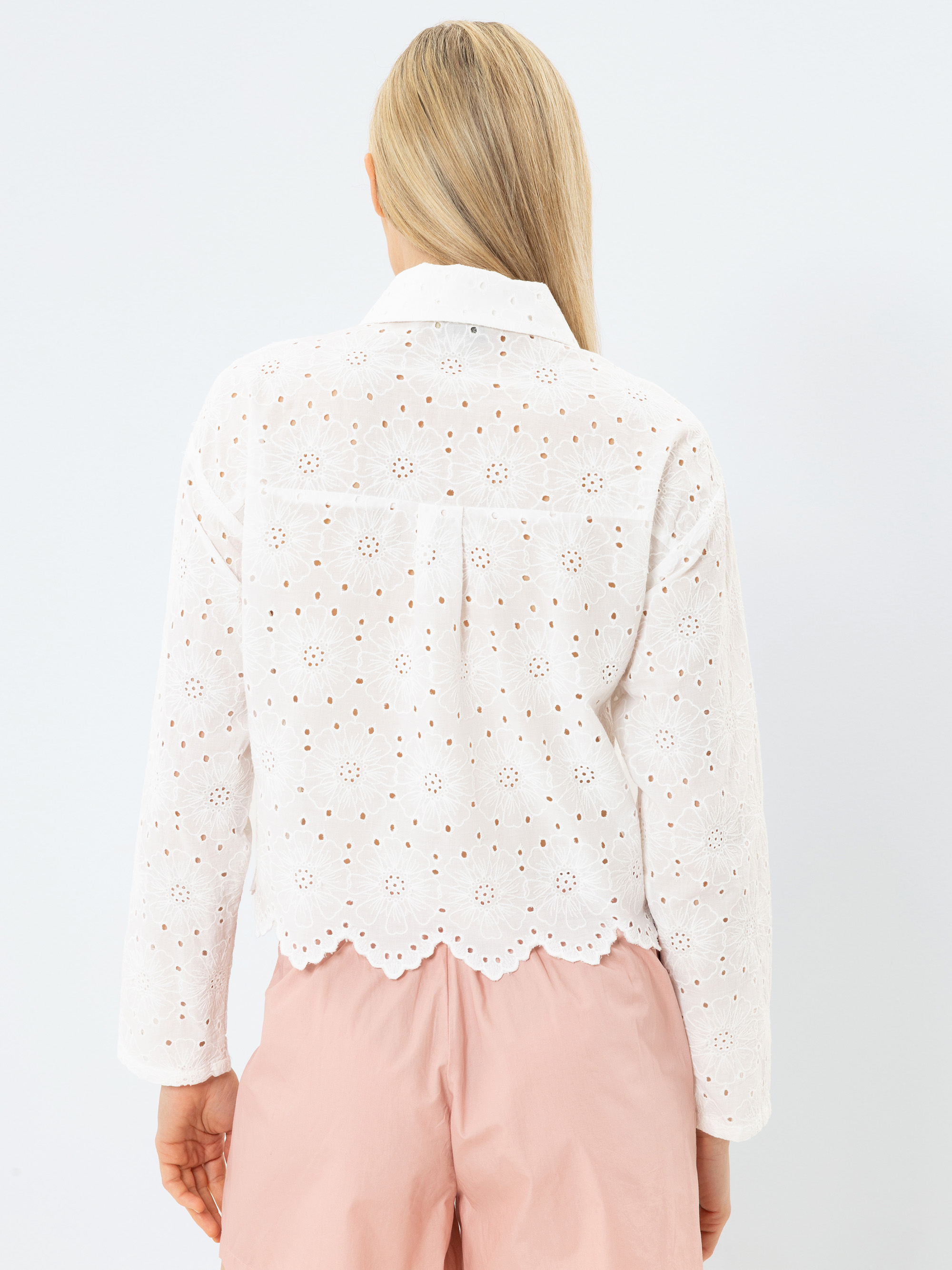 Eine Person mit langen blonden Haaren ist von hinten zu sehen. Sie trägt eine weiße, langärmelige Ösenbluse mit Blumenmustern und Wellenkanten, gepaart mit hellrosa Shorts vor einem schlichten hellen Hintergrund.