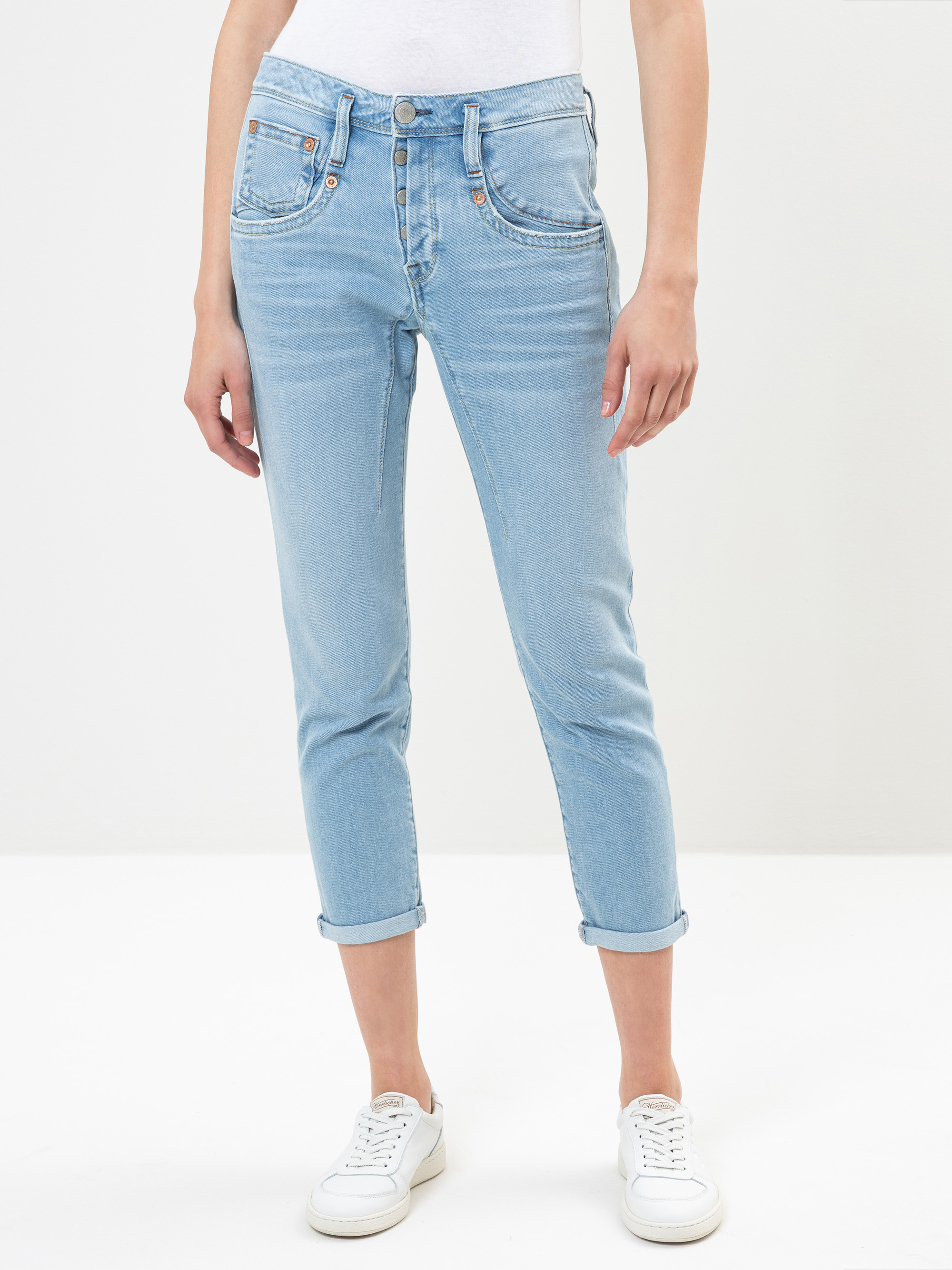 Person in hellblauer Jeans mit Bündchen, weißem Oberteil und weißen Turnschuhen vor einem schlichten weißen Hintergrund. Die Jeans hat eine leicht lockere Passform und sichtbare Taschen.