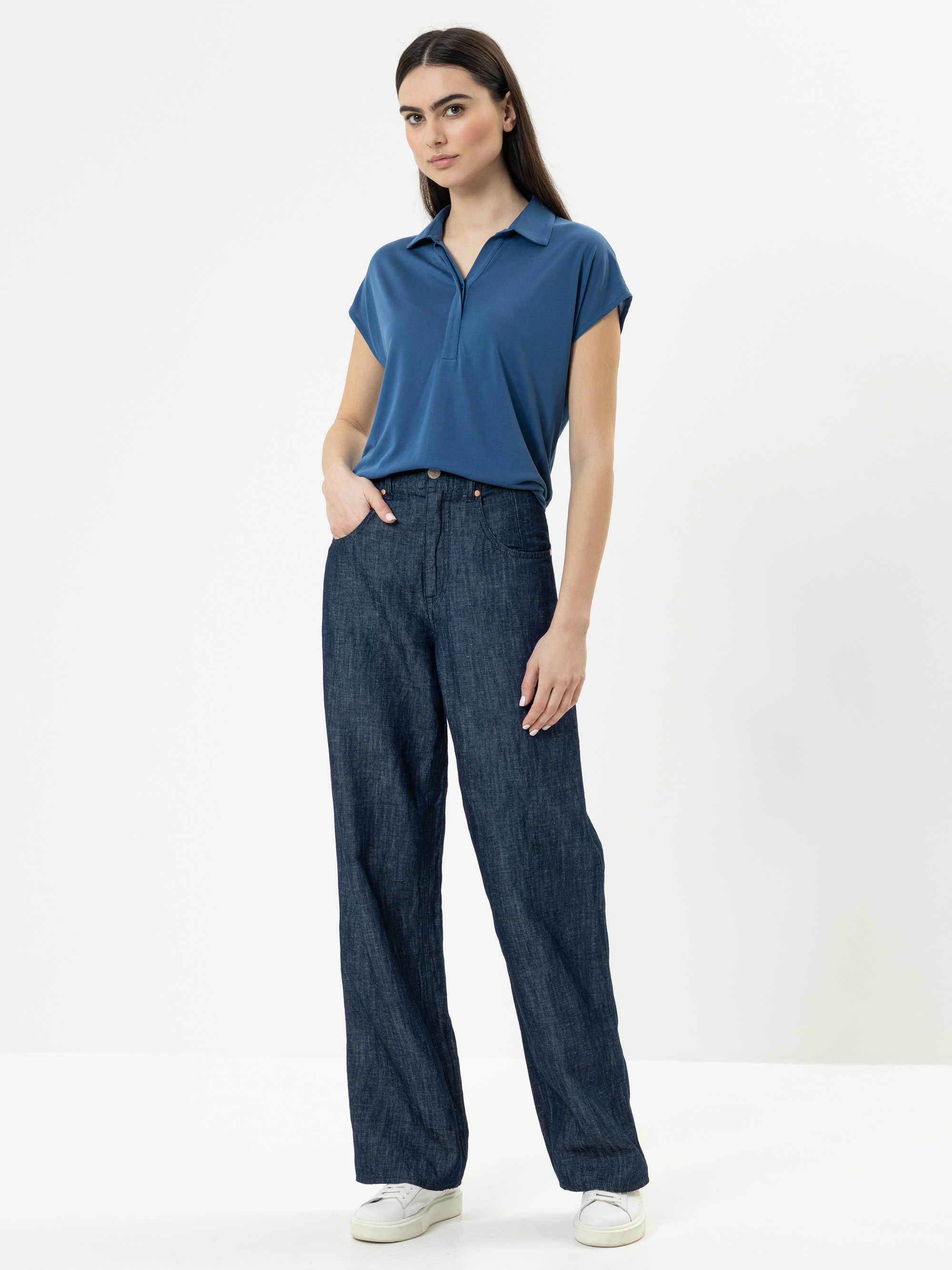 Eine Frau steht vor einem schlichten weißen Hintergrund und trägt ein kurzärmeliges blaues Poloshirt, eine hochtaillierte Jeans mit weitem Bein und weiße Turnschuhe. Sie blickt mit einer Hand in der Tasche nach vorne.