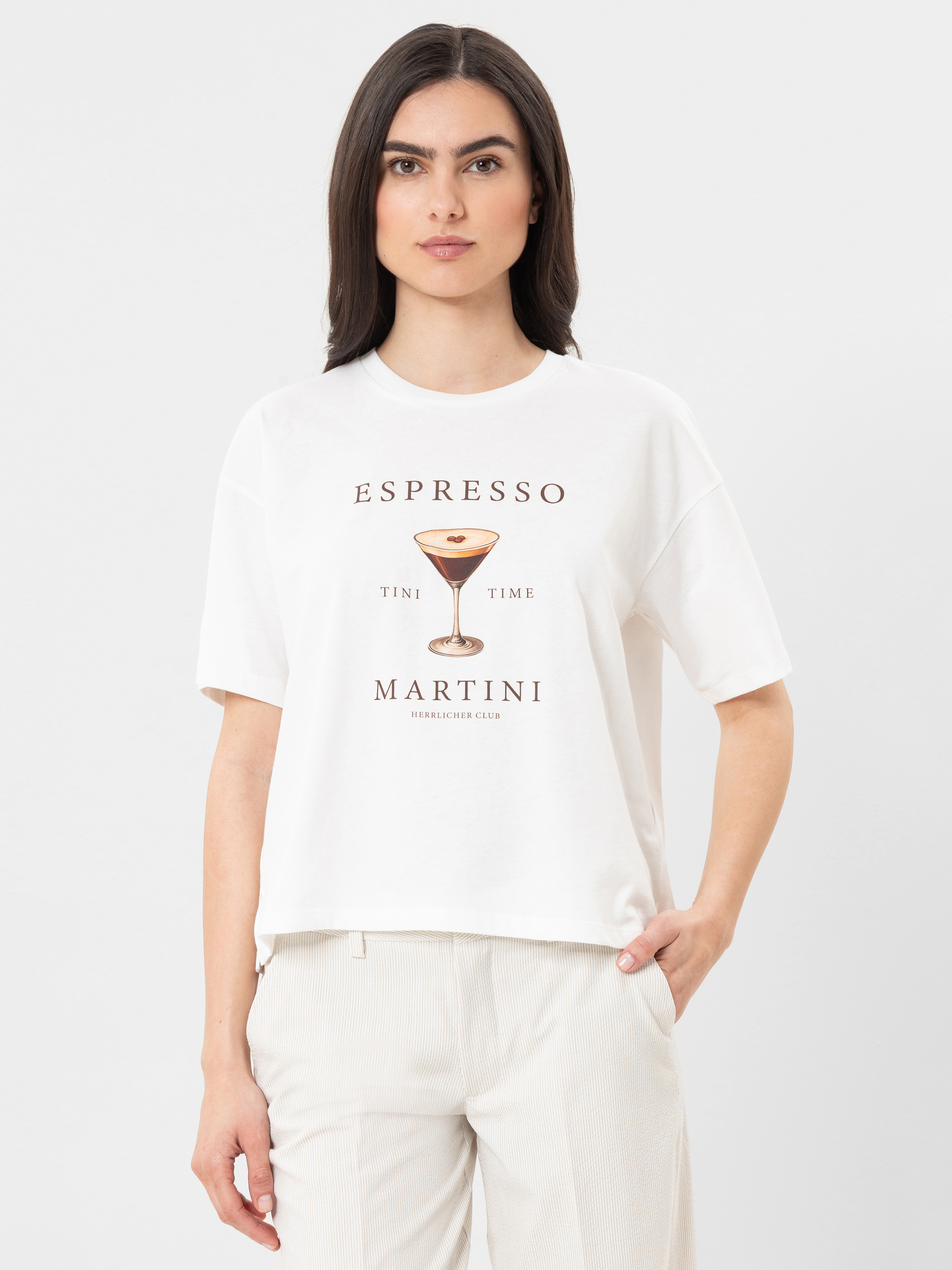 Eine Frau mit glattem braunem Haar, die ein weißes T-Shirt mit der Abbildung eines Martiniglases und der Aufschrift "ESPRESSO MARTINI TIME" trägt, steht mit einer Hand in der Tasche vor einem unifarbenen Hintergrund.