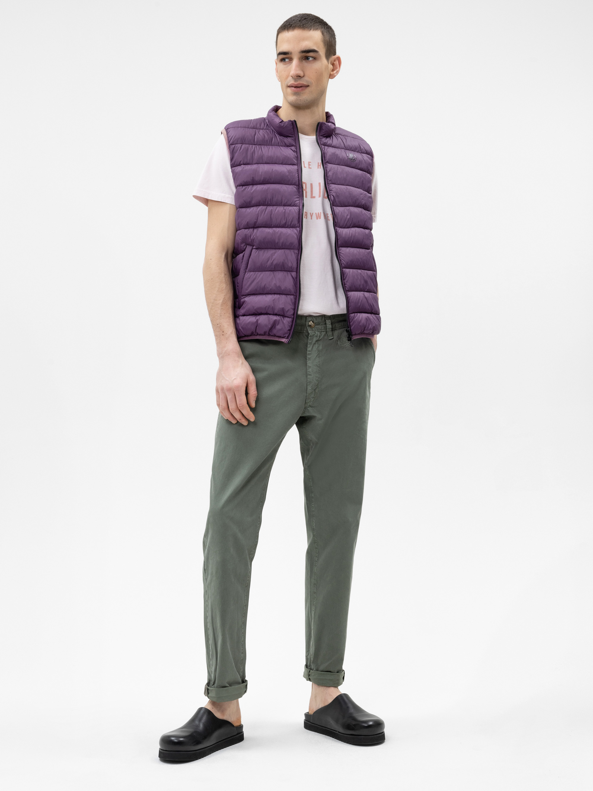 Eine Person steht vor einem schlichten weißen Hintergrund und trägt eine lila Pufferweste, ein hellrosa grafisches T-Shirt, eine olivgrüne Hose und schwarze Slipper.