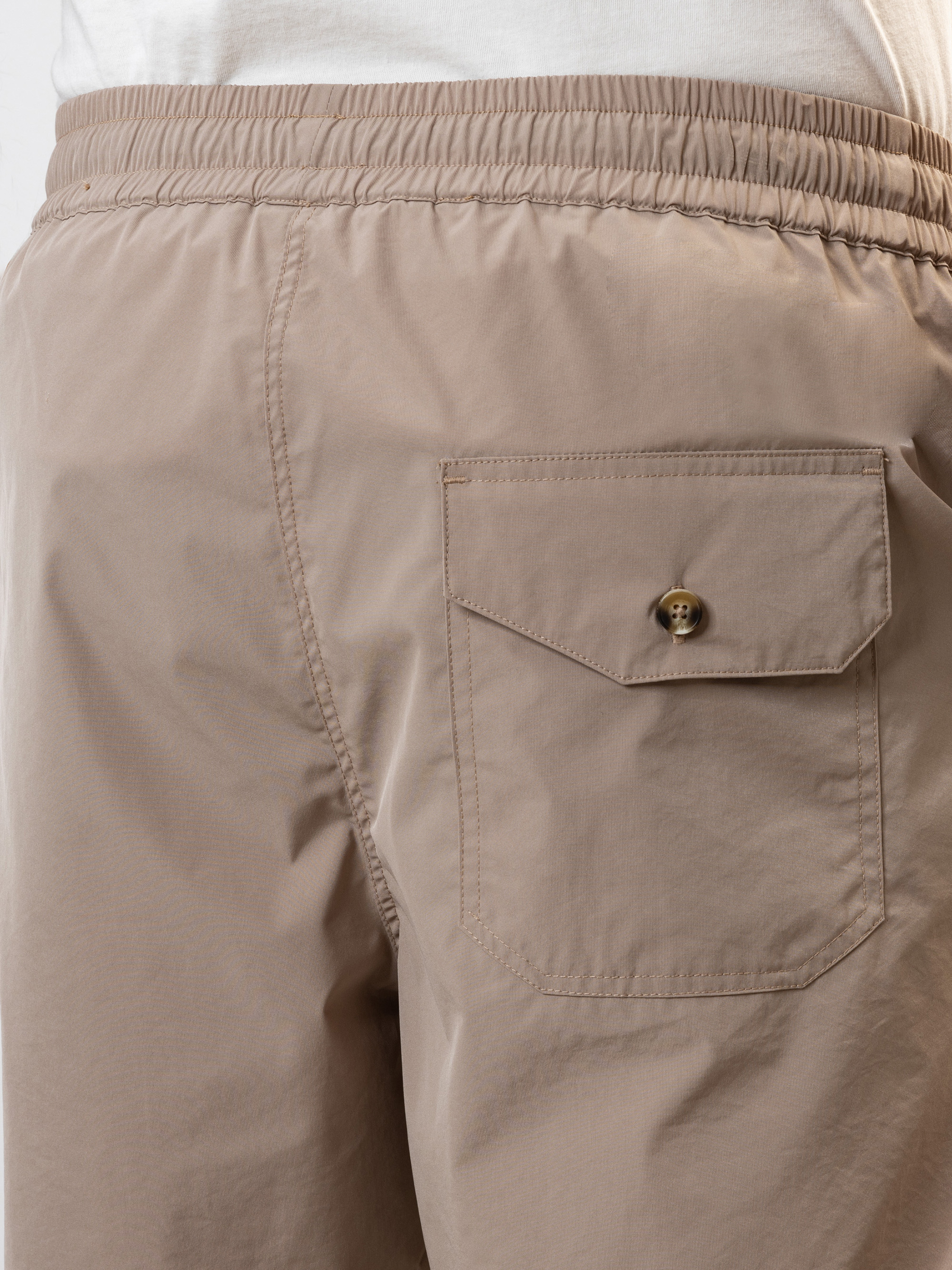 Nahaufnahme des Rückens einer Person, die eine hellbeige Hose mit elastischer Taille trägt, die auf der rechten Seite eine Klappentasche mit Knopf aufweist. Die Person trägt außerdem ein weißes Hemd.