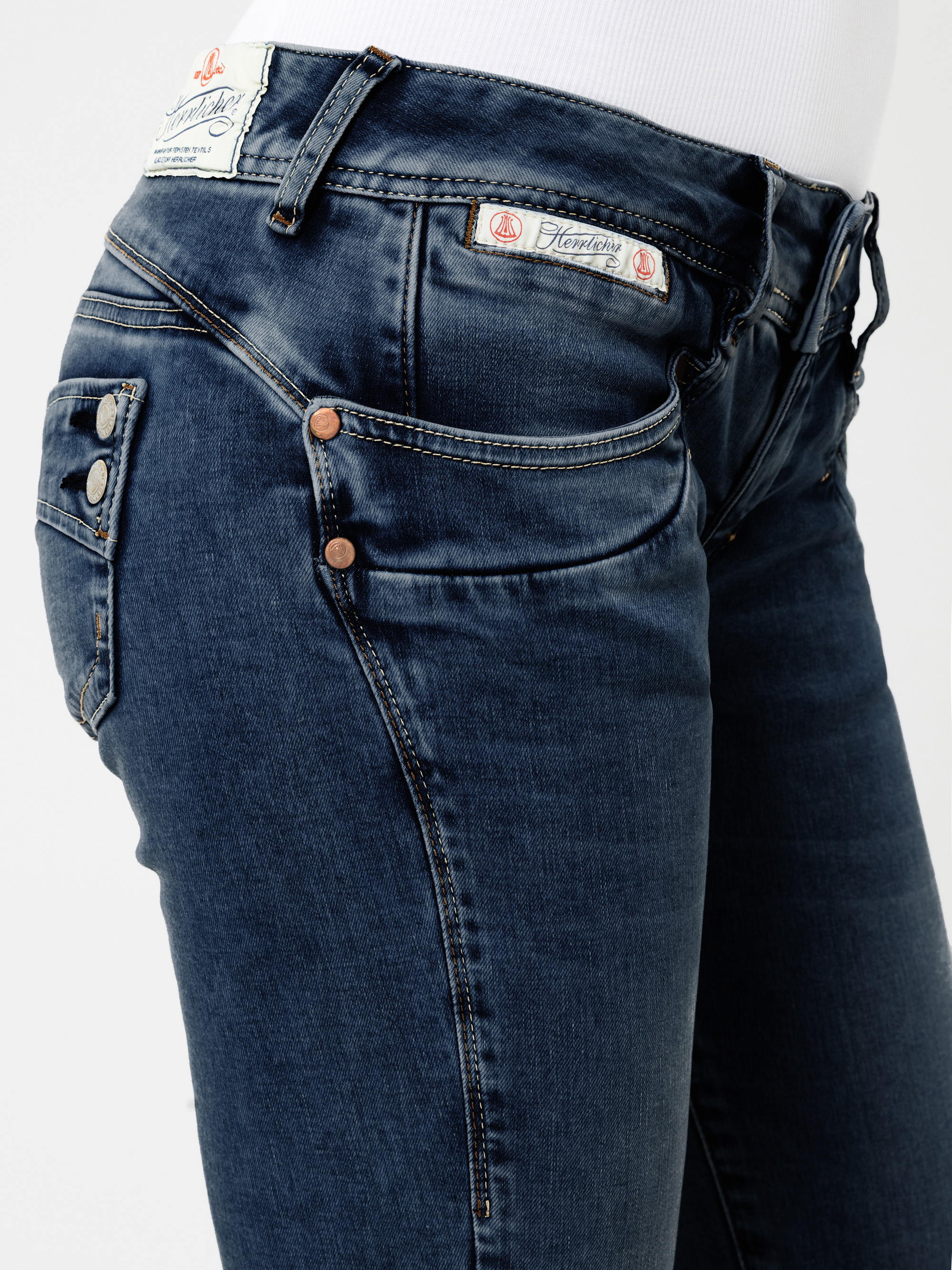 Nahaufnahme einer Person, die eine dunkelblaue Jeans mit kupferfarbenen Nieten, Ziernähten und zwei Logo-Aufnähern am Bund trägt, dazu ein weißes Oberteil. Zu sehen sind nur der untere Rumpf und die Oberschenkel.