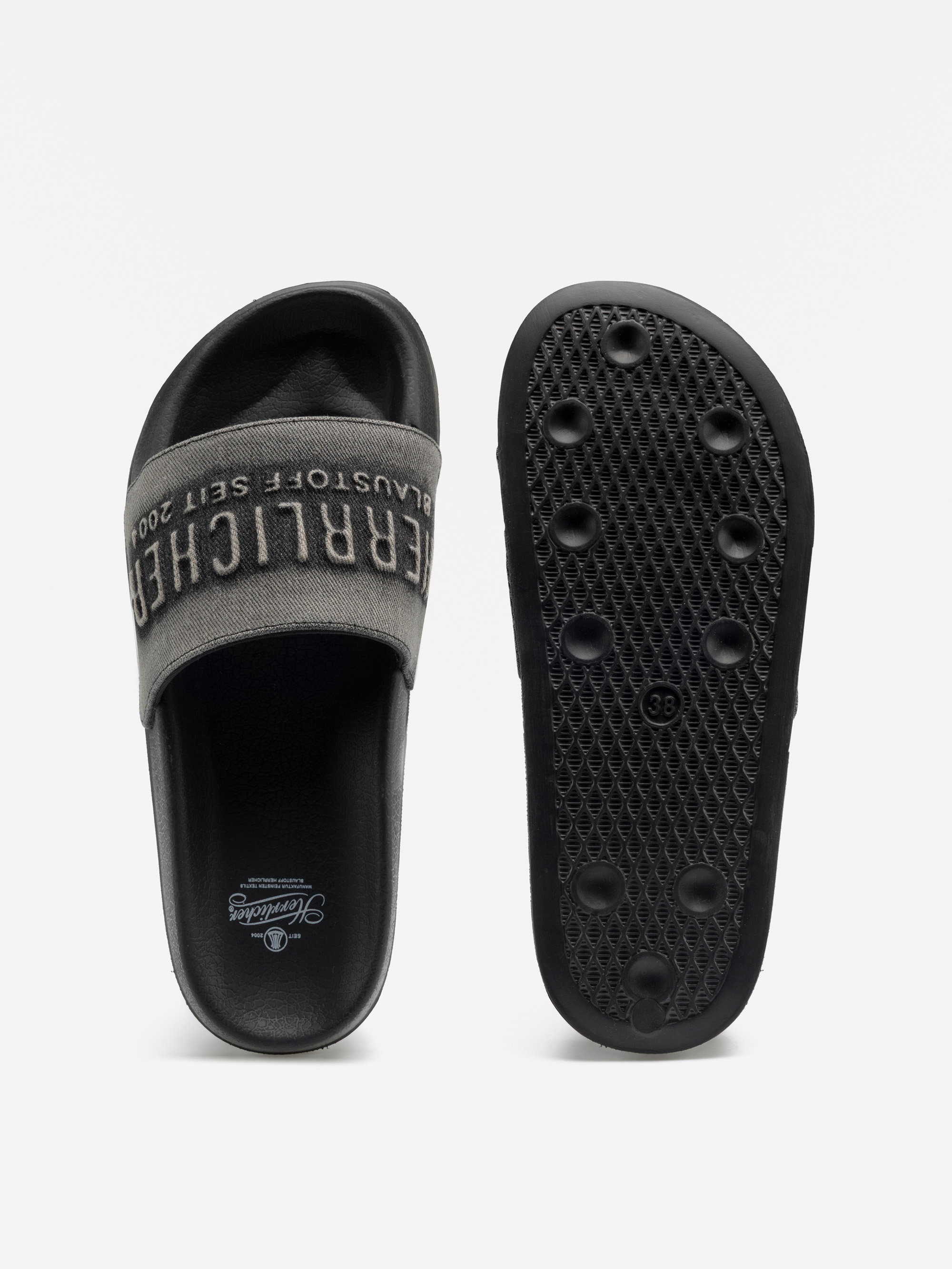 Zwei schwarze Slide-Sandalen, eine oben mit einem breiten grauen Riemen, auf dem Text aufgedruckt ist, die andere mit einer strukturierten Sohle mit rundem Griffmuster. Der Markenname ist auf der Innensohle sichtbar.