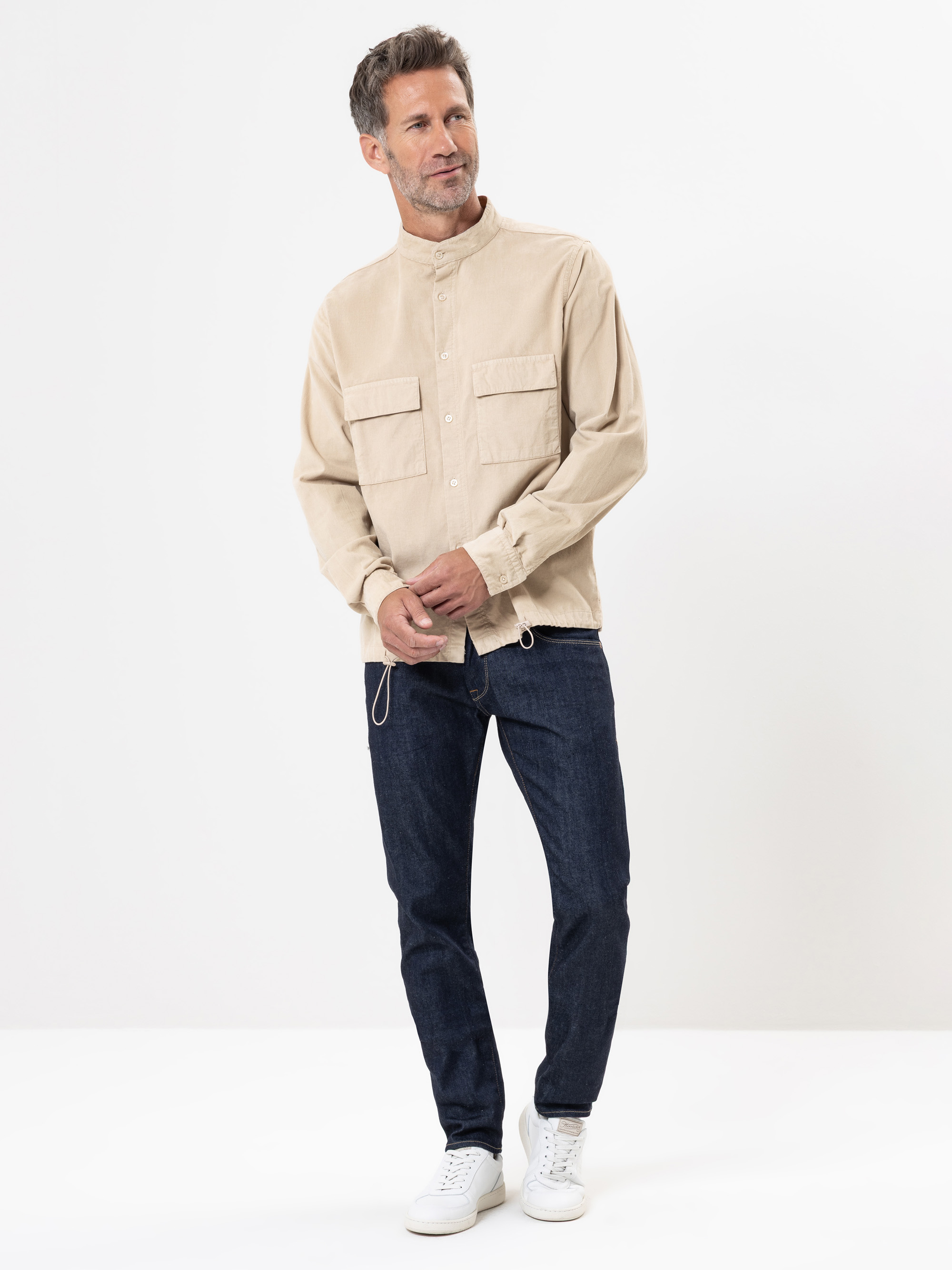 Ein Mann steht vor einem weißen Hintergrund und trägt ein beiges Button-up-Hemd mit zwei Brusttaschen, dunkelblaue Jeans und weiße Turnschuhe. Er blickt leicht zur Seite und hat die Hände vor sich verschränkt.
