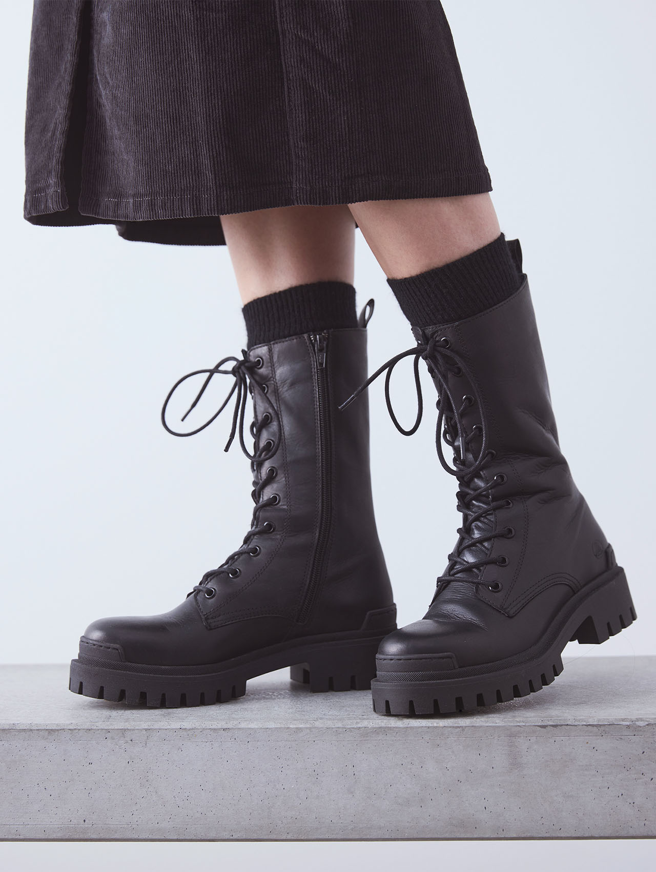 Eine Person, die schwarze Schnürstiefel mit dicken Sohlen und schwarze Socken trägt, steht auf einer grauen Fläche. Die Person trägt außerdem einen dunklen Rock. Der Hintergrund ist einfarbig und hell gefärbt.
