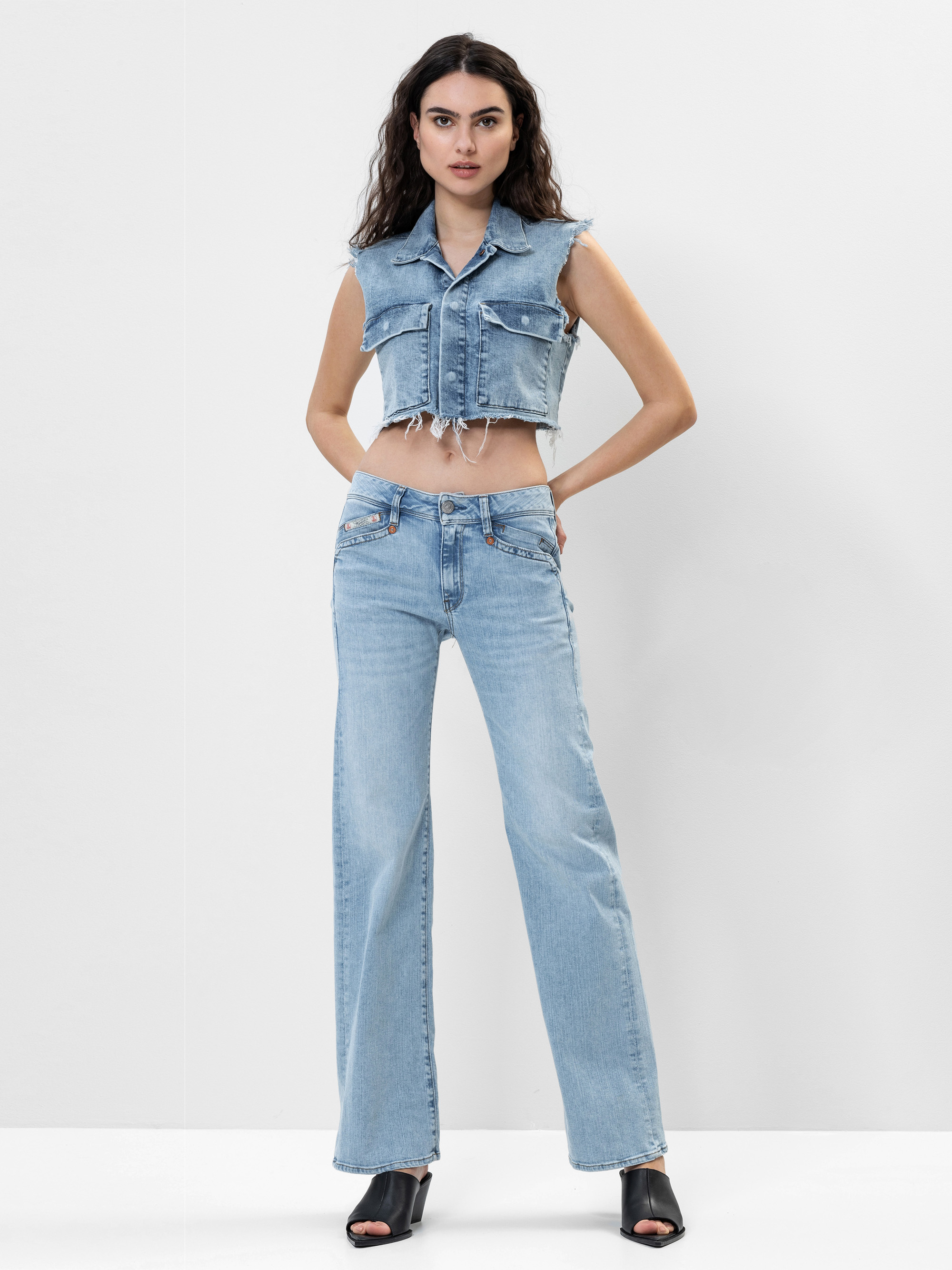 Eine Frau steht vor einem einfarbig weißen Hintergrund und trägt eine ärmellose, kurzgeschnittene Jeansweste und hellblaue Jeans mit hoher Taille und weiten Beinen, gepaart mit schwarzen, offenen Absätzen.