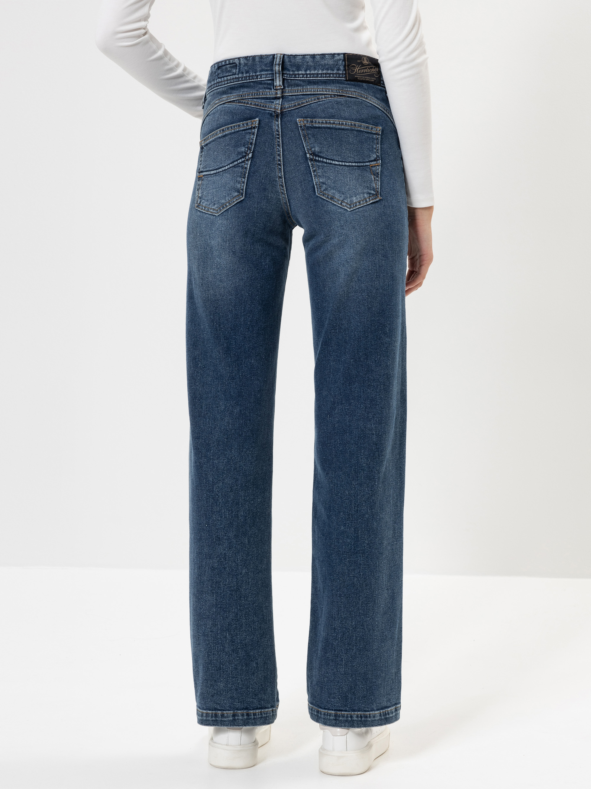 Eine Person, die eine blaue Jeans mit hoher Taille und weitem Bein sowie weiße Turnschuhe trägt, wird von hinten vor einem schlichten weißen Hintergrund gezeigt. Die Person trägt außerdem ein langärmeliges weißes Oberteil.