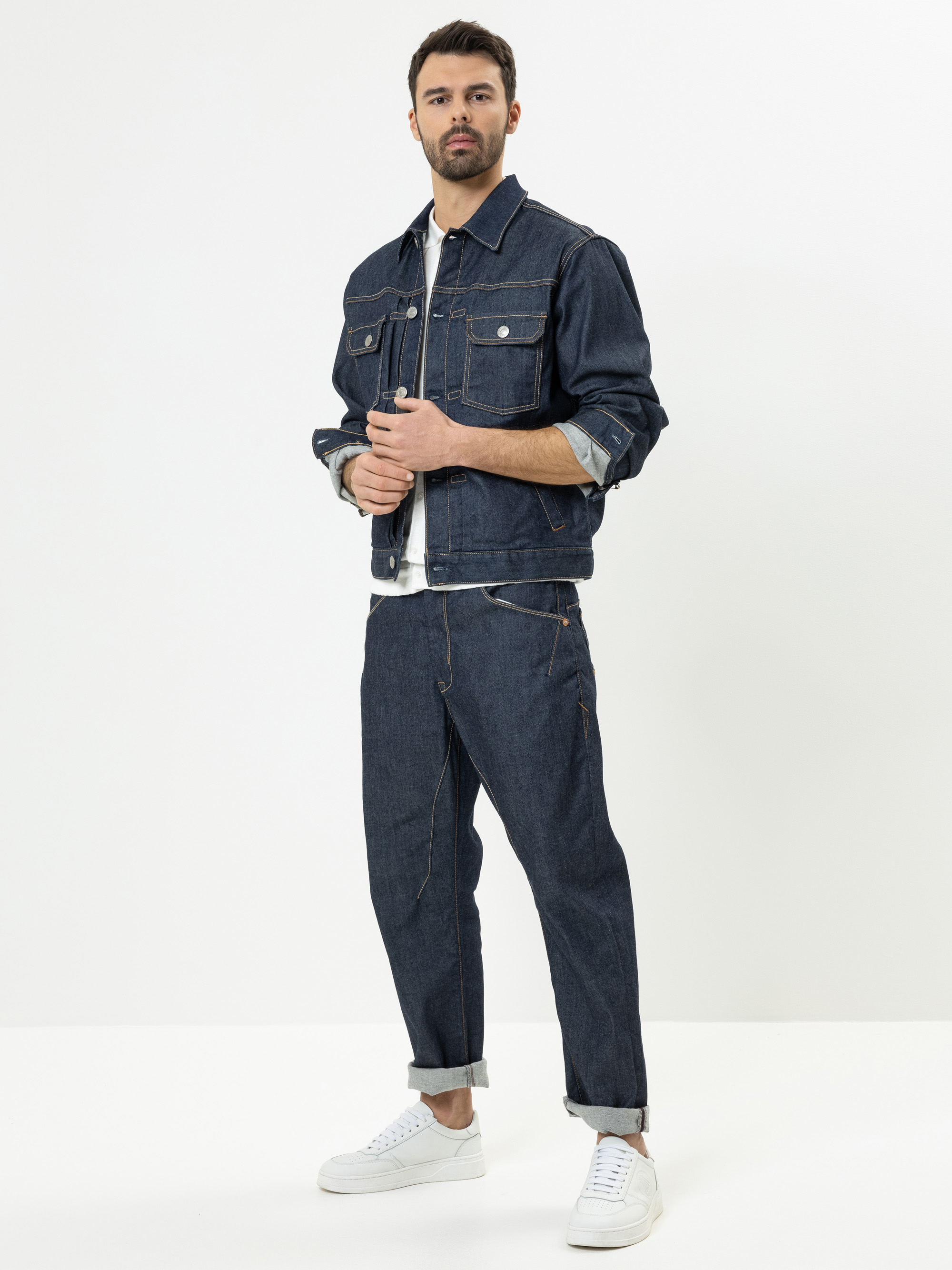 Ein Mann steht vor einem weißen Hintergrund und trägt eine dunkelblaue Jeansjacke und eine dazu passende Jeans, ein weißes T-Shirt und weiße Turnschuhe. Mit den Händen rückt er die Vorderseite seiner Jacke zurecht und blickt mit neutraler Miene nach