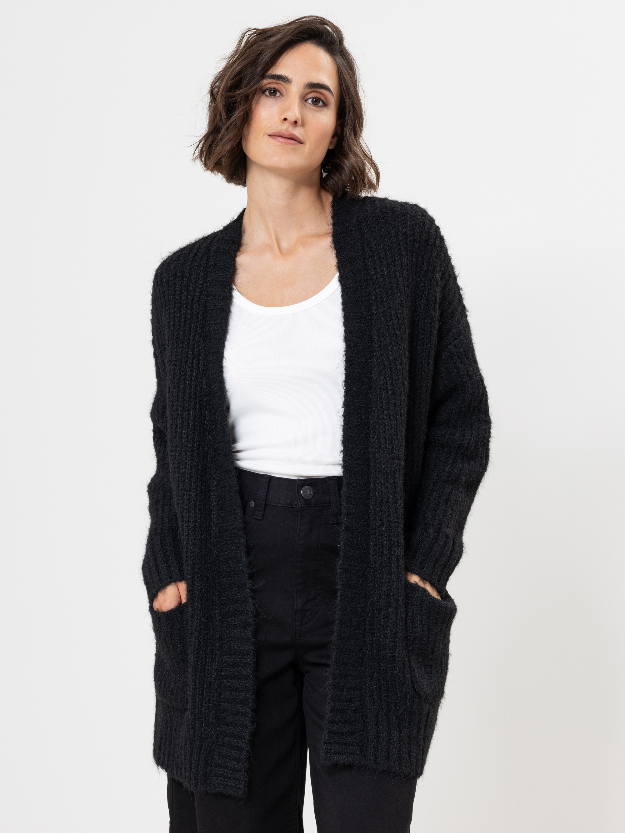 Eine Frau mit kurzen braunen Haaren trägt ein weißes Tanktop, eine schwarze Strickjacke und eine schwarze Hose. Sie steht vor einem schlichten weißen Hintergrund und hat die Hände in den Taschen ihrer Strickjacke.