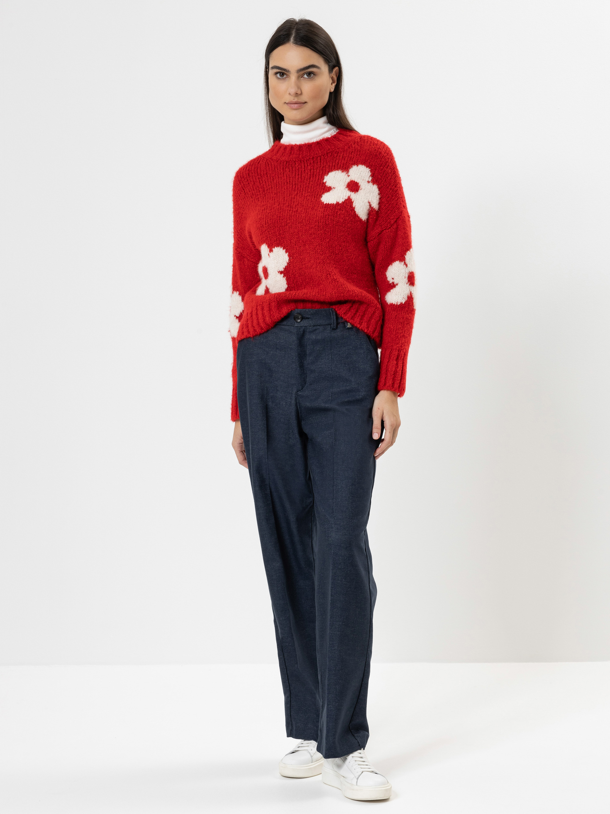 Eine Frau steht vor einem weißen Hintergrund und trägt einen roten Pullover mit weißen Blumenmustern, eine blaue Hose, weiße Turnschuhe und darunter einen weißen Rollkragenpullover. Sie hat langes dunkles Haar und einen neutralen Gesichtsausdruck.