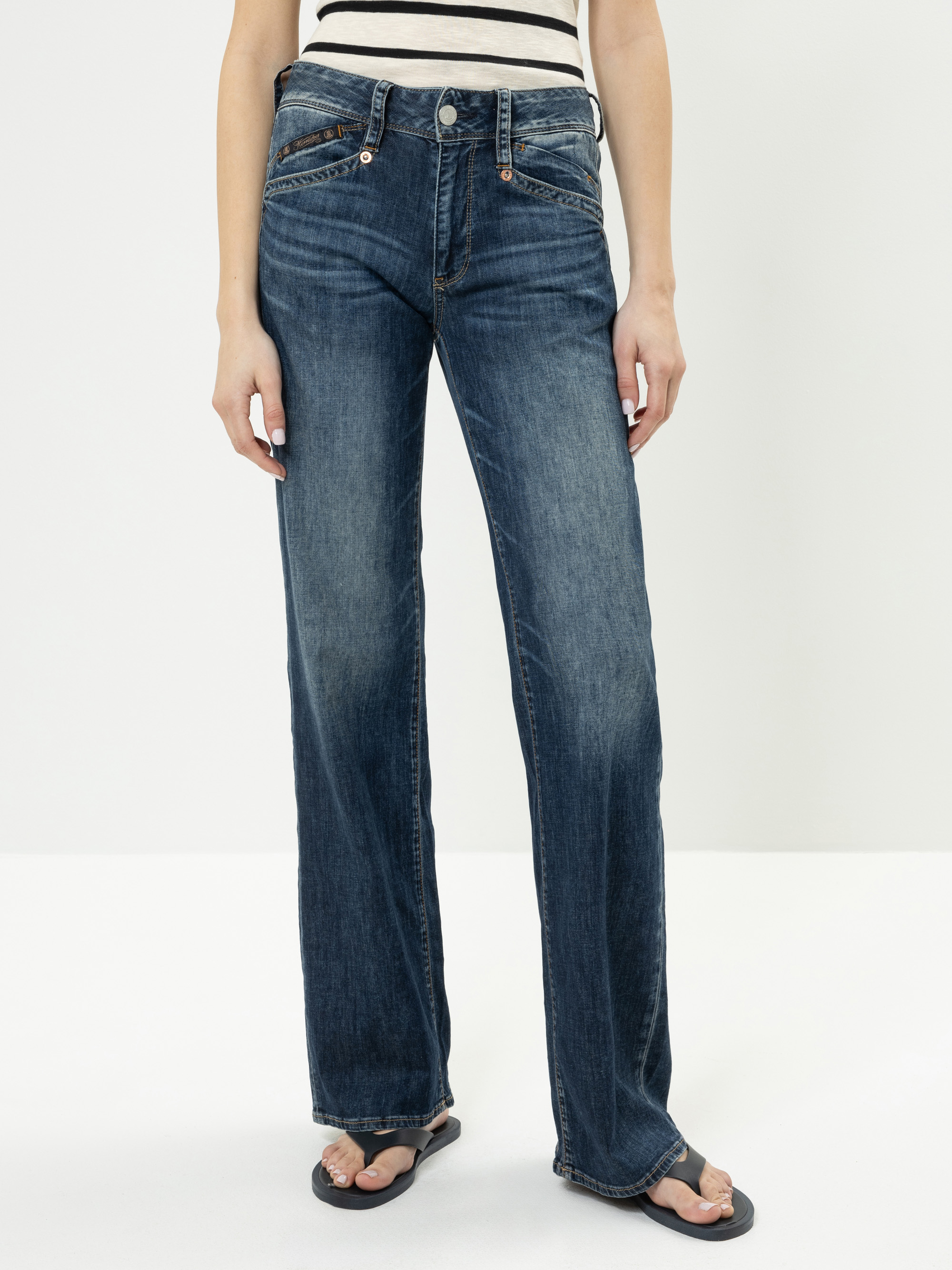 Eine Person, die eine blaue Jeans mit hoher Taille und geradem Bein mit verblasster Waschung, ein weißes Oberteil mit schwarzen Streifen und schwarze Flip-Flops trägt, steht vor einem schlichten weißen Hintergrund.