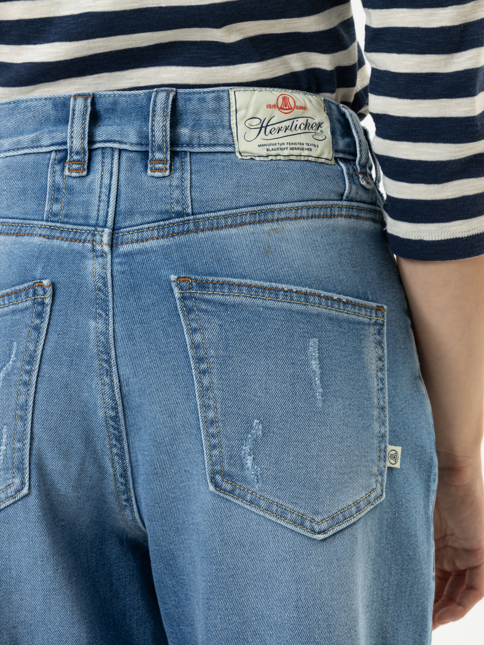 Nahaufnahme einer Person, die hellblaue Jeans und ein marineblau-weiß gestreiftes Hemd trägt. Die Jeans hat ein weißes Etikett am Bund und sichtbare Gesäßtaschen mit dezenten Used-Elementen.
