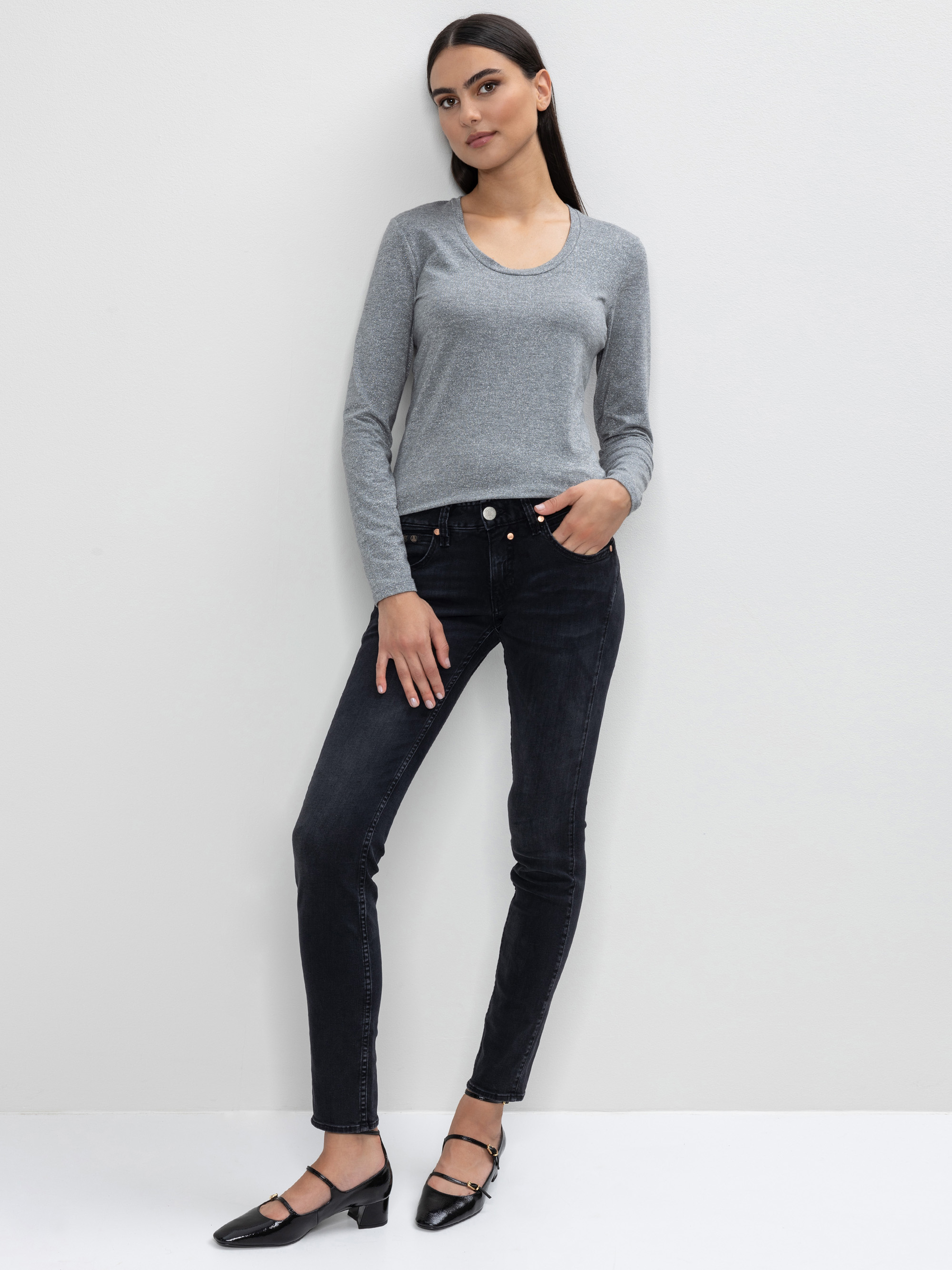 Eine Frau steht vor einer weißen Wand und trägt ein graues langärmeliges Oberteil, schwarze Jeans und schwarze Riemenschuhe. Sie hat langes dunkles Haar und ihre Arme liegen entspannt an ihren Seiten, ein Bein ist leicht angewinkelt.