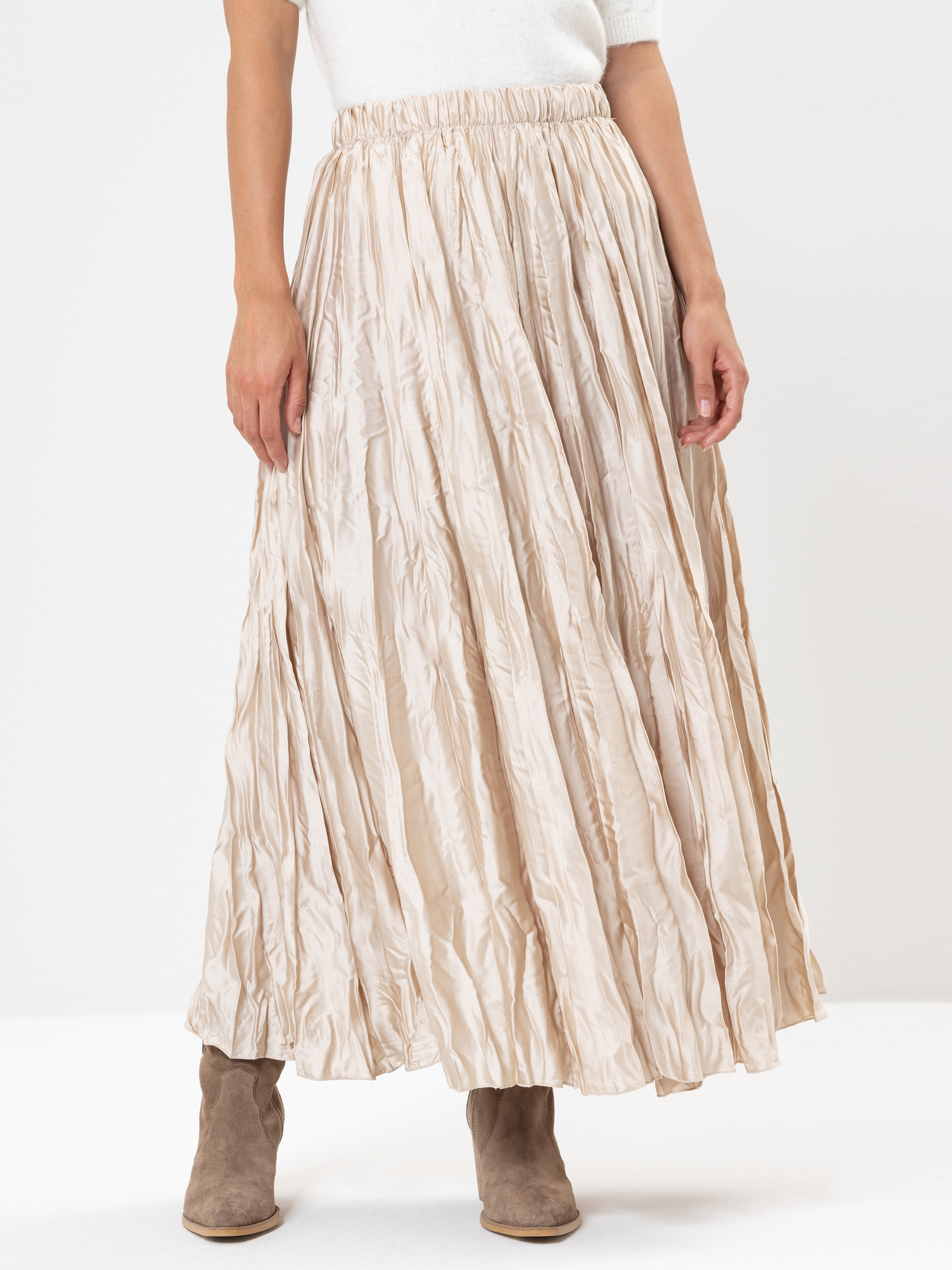 Eine Person, die ein weißes Oberteil und einen langen beigefarbenen Faltenrock mit Knitterstruktur trägt, gepaart mit hellbraunen Wildlederstiefeln, steht vor einem schlichten weißen Hintergrund.