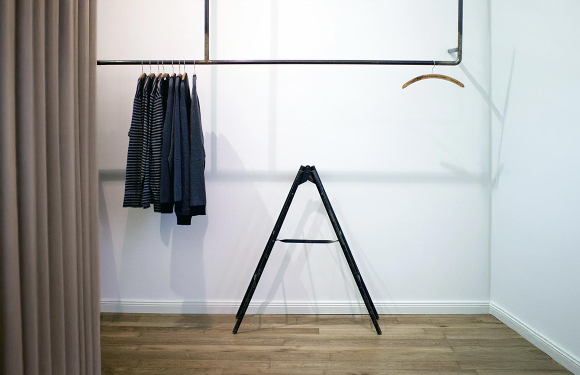 Ein minimaler Kleiderschrank mit vier gestreiften und einfarbigen Jacken, die auf einem schwarzen Regal hängen, einem leeren Holzbügel, einer kleinen schwarzen Trittleiter und einem beigen Vorhang auf der linken Seite, alles vor einer weißen Wand und e