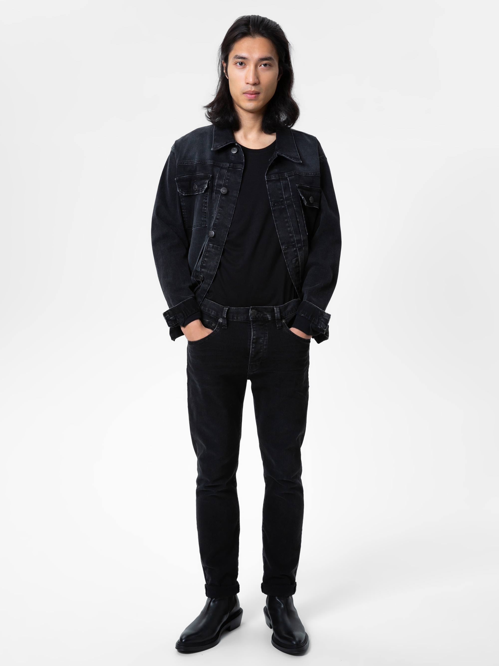 Eine Person mit langen dunklen Haaren steht mit dem Gesicht nach vorne und trägt eine schwarze Jeansjacke, ein schwarzes T-Shirt, schwarze Jeans und schwarze Stiefel vor einem schlichten weißen Hintergrund. Ihre Hände stecken in den Taschen.