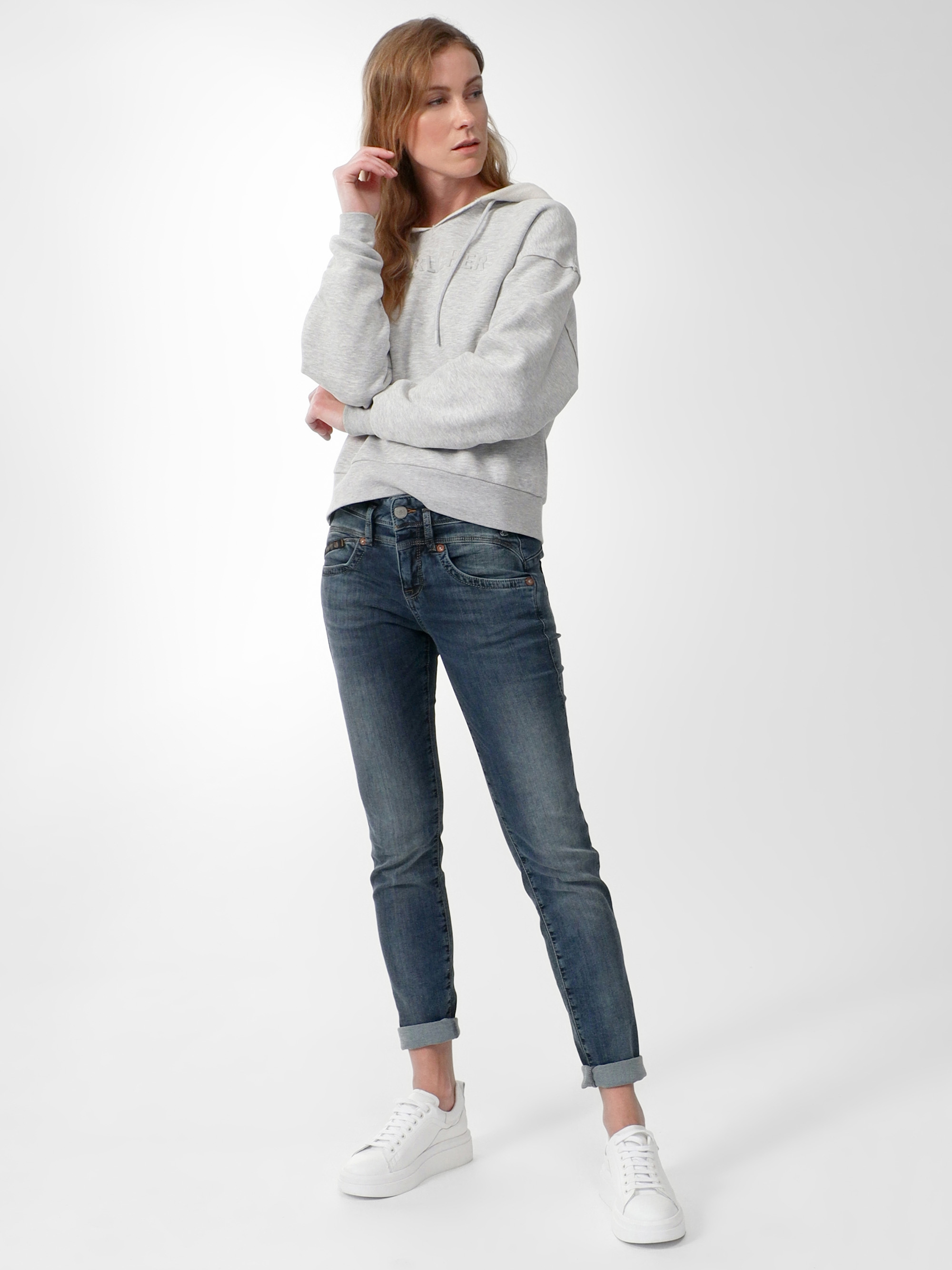 Eine Person steht vor einem schlichten Hintergrund, trägt einen hellgrauen Kapuzenpullover, Jeans mit blauen Bündchen und weiße Turnschuhe, eine Hand vor dem Gesicht und einen nachdenklichen Gesichtsausdruck.