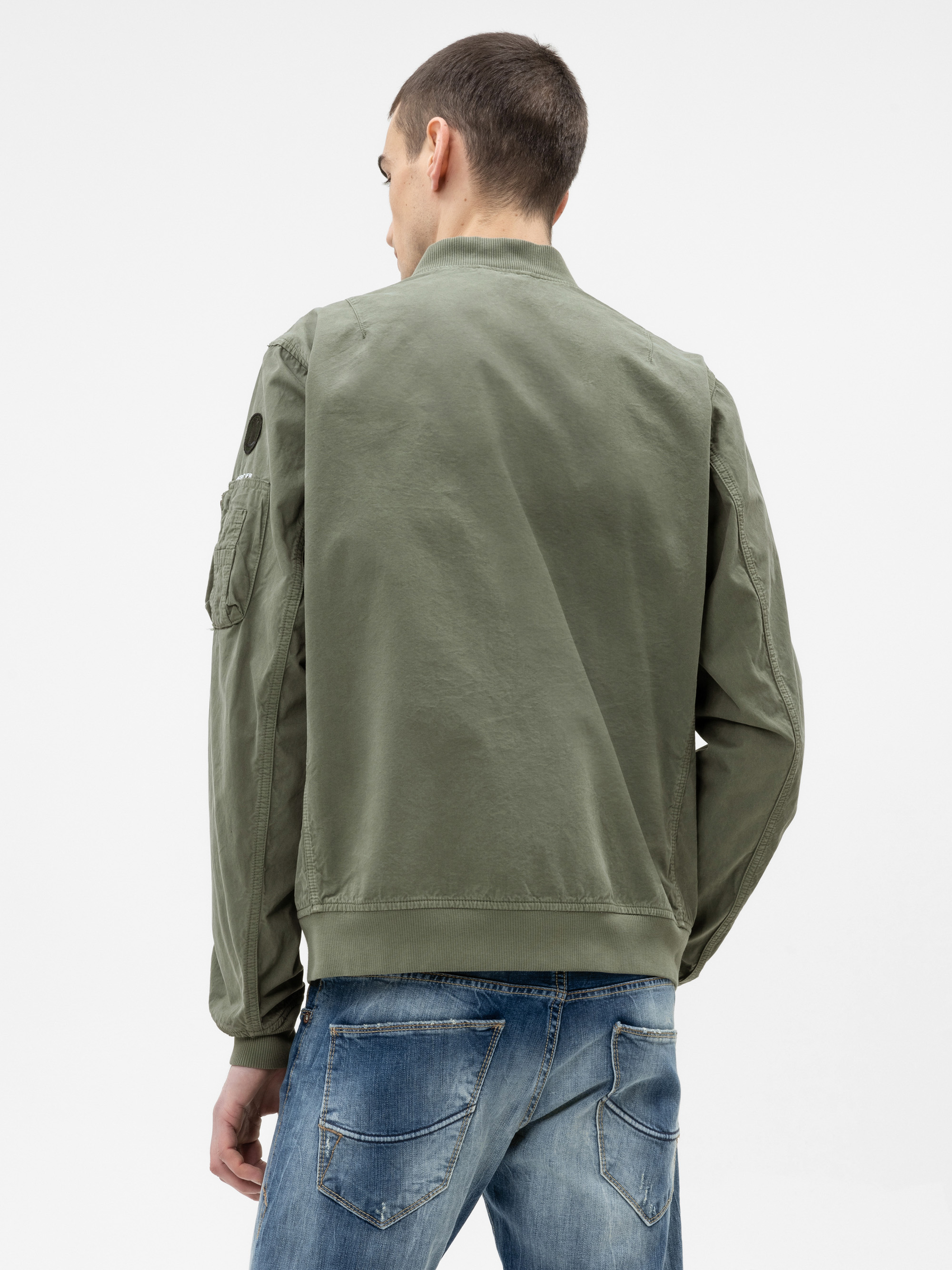 Eine Person, die eine olivgrüne Jacke und blaue Jeans trägt, steht mit dem Rücken zur Kamera vor einem schlichten weißen Hintergrund.