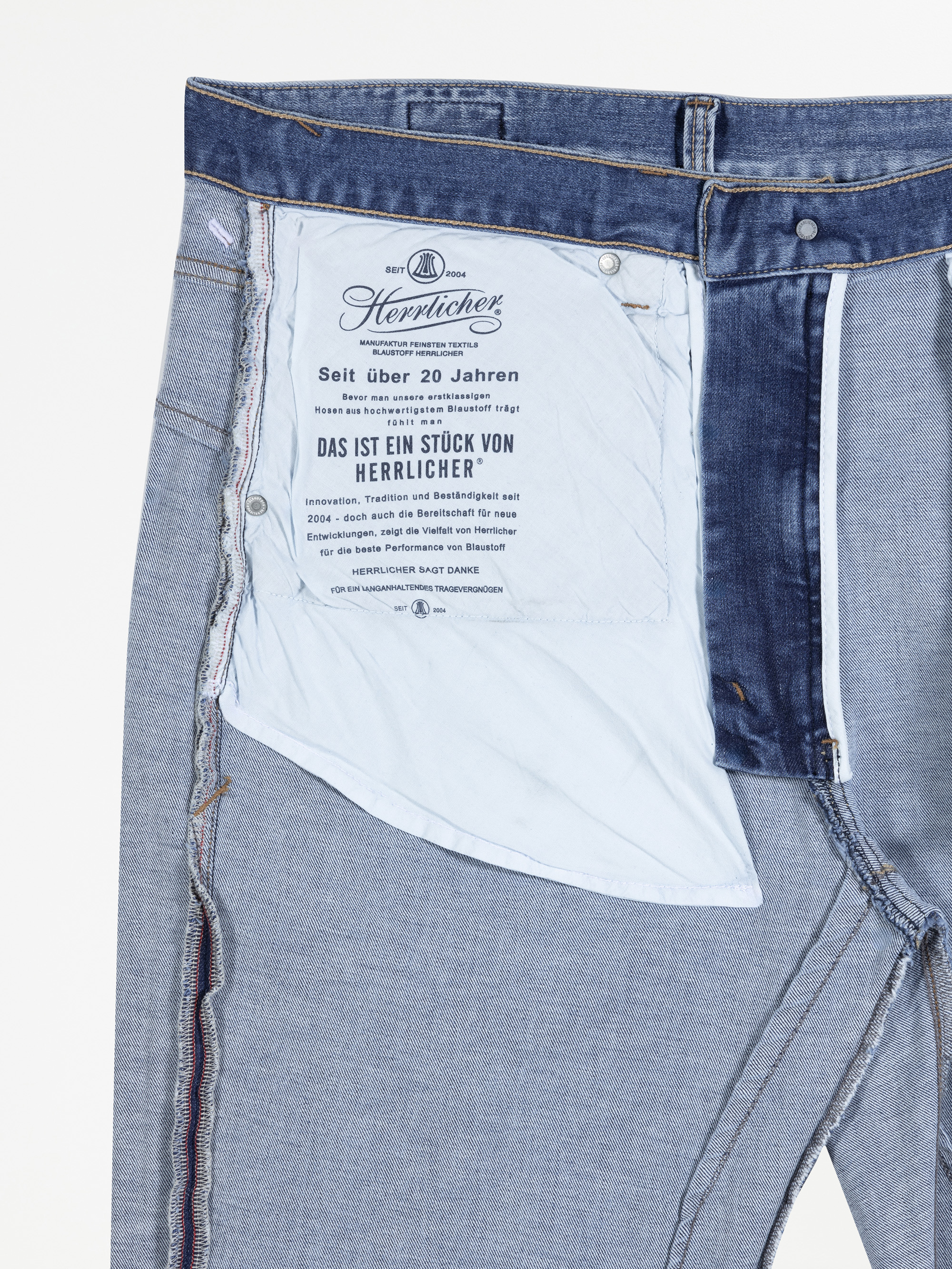 Nahaufnahme der Innenseite einer blauen Jeans, die das Innentaschenfutter mit aufgedrucktem Text und Branding der Firma Herrlicher zeigt. Die Jeans ist teilweise aufgeknöpft und liegt flach.