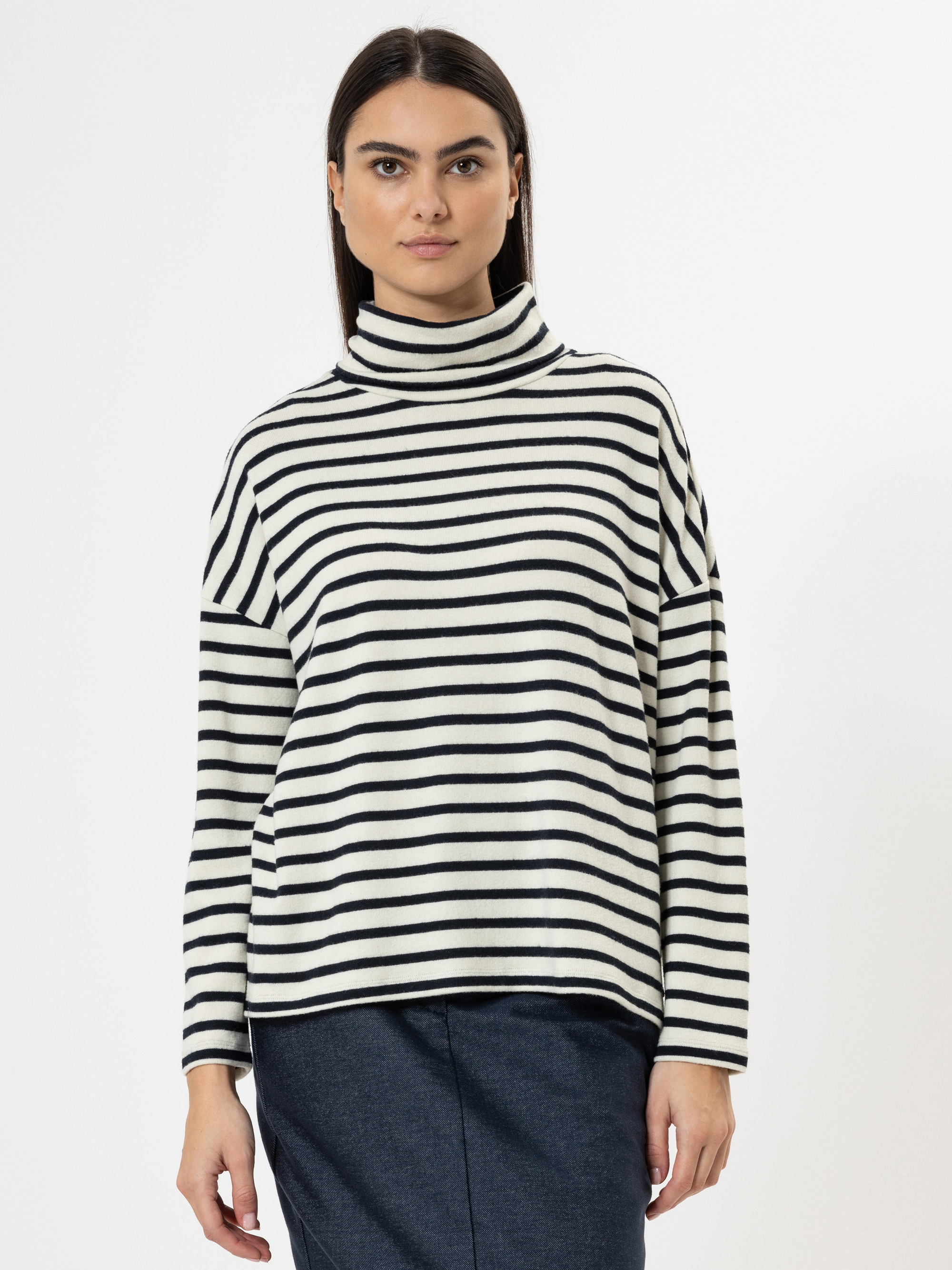 Eine Frau mit glattem dunklem Haar trägt einen schwarz-weiß gestreiften Rollkragenpullover und eine dunkle Hose und steht vor einem schlichten weißen Hintergrund.