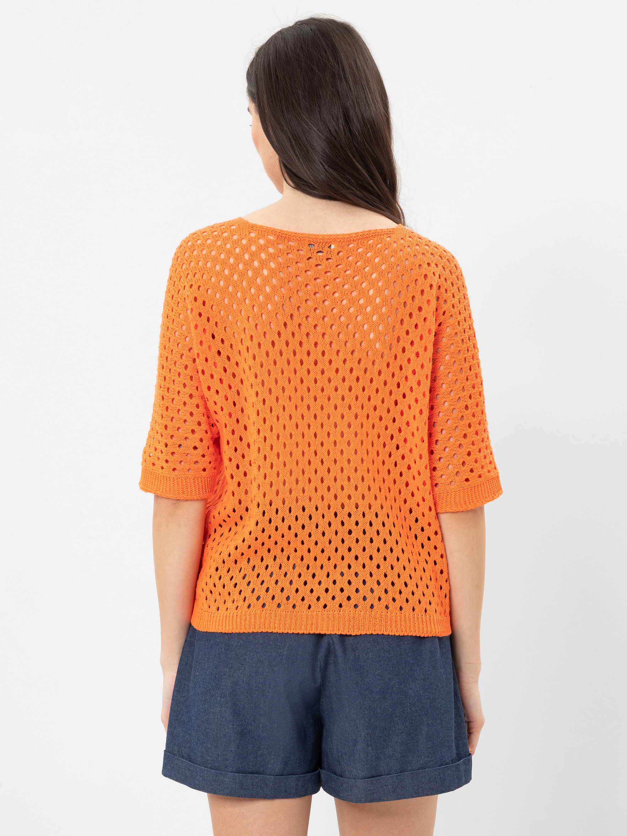 Eine Frau mit langen braunen Haaren, von hinten gesehen, trägt einen orangefarbenen, kurzärmeligen, locker gestrickten Pullover und dunkelblaue Shorts vor einem schlichten weißen Hintergrund.