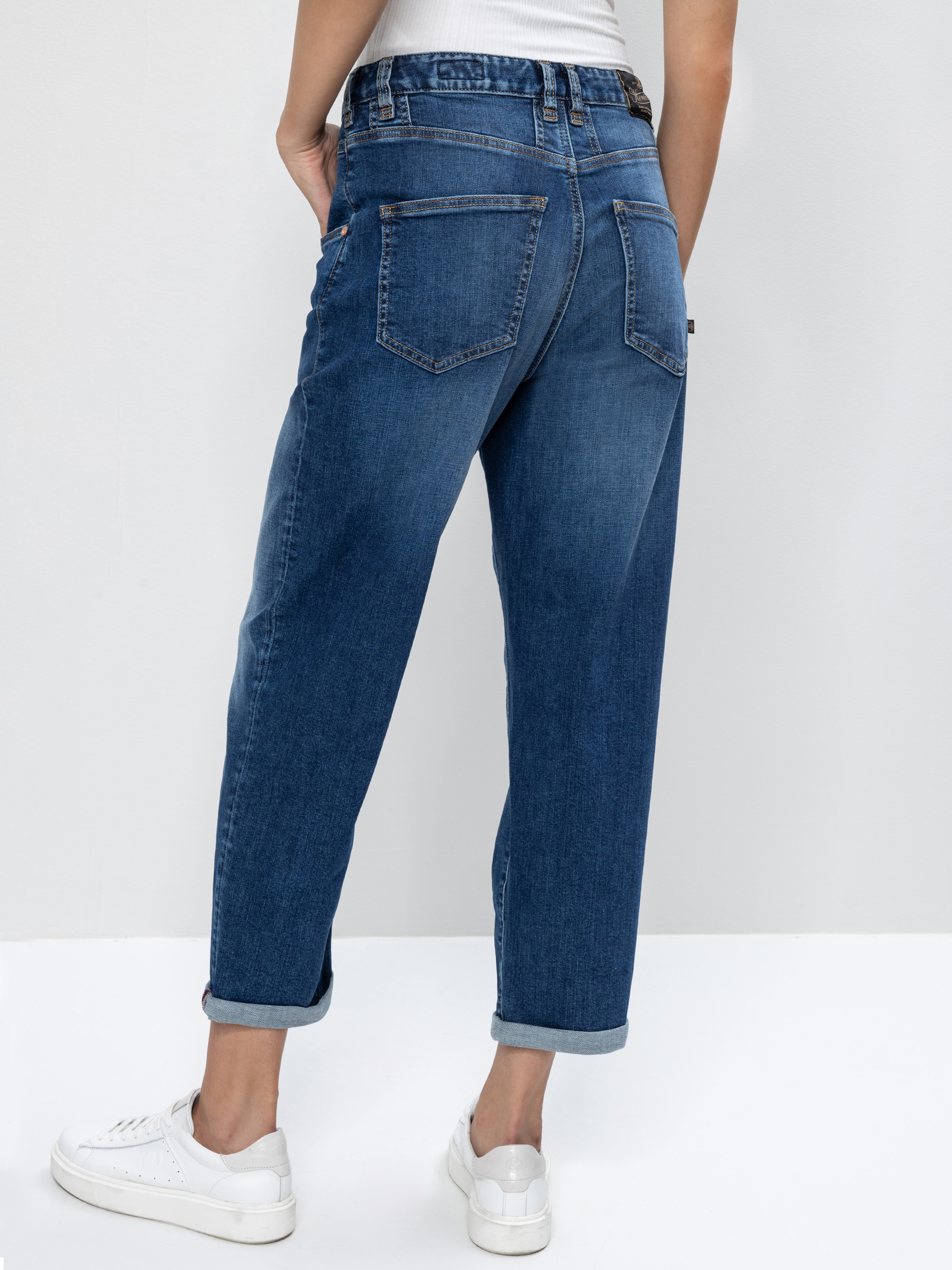 Eine Person steht mit dem Gesicht zur Wand und trägt eine blaue Jeans mit Bündchen, ein weißes ärmelloses Oberteil und weiße Turnschuhe vor einem schlichten hellen Hintergrund.