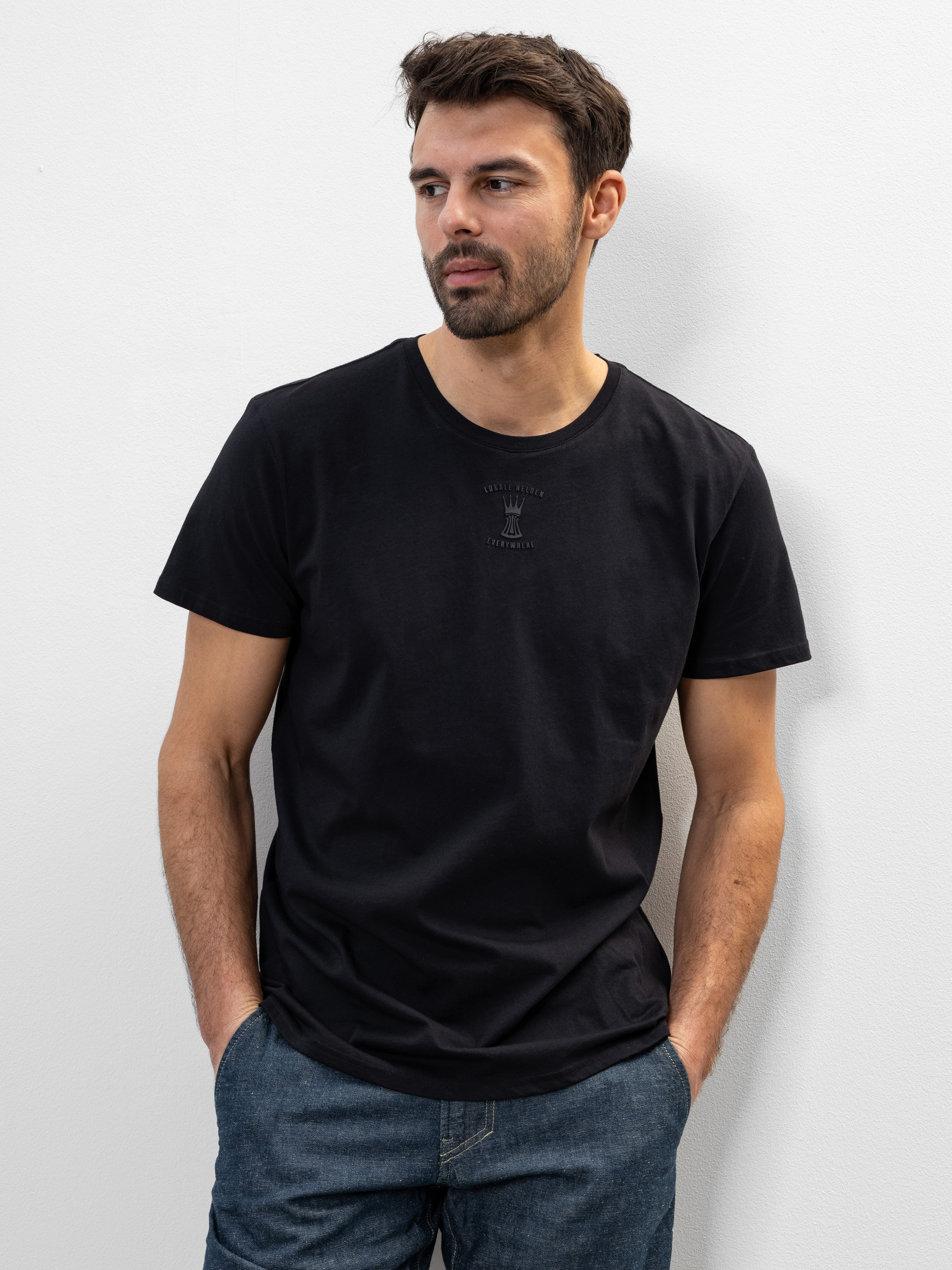 Ein Mann mit kurzen dunklen Haaren und einem Bart trägt ein schwarzes T-Shirt und blaue Jeans. Er steht an einer schlichten weißen Wand, schaut leicht zur Seite und hat die Hände in den Taschen.