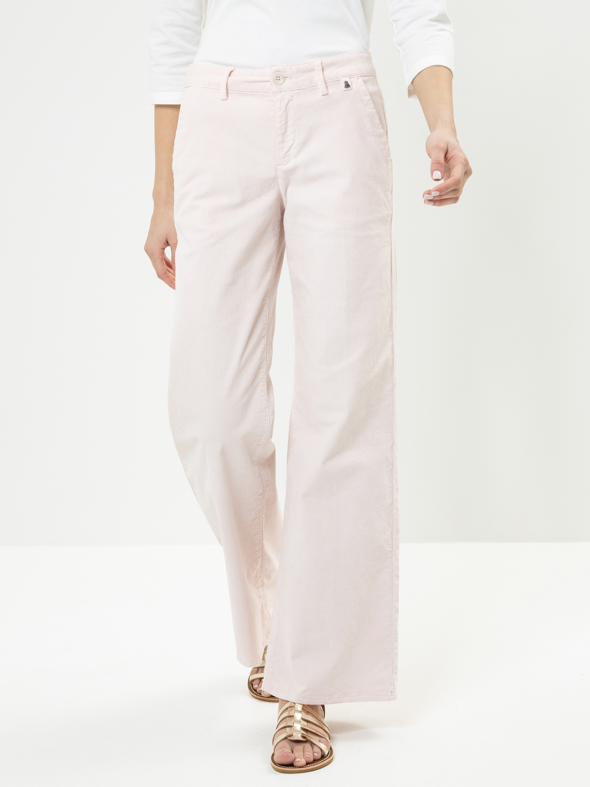 Eine Person, die eine hellrosa Hose mit weitem Bein, ein weißes langärmeliges Oberteil und goldene Sandalen trägt, steht vor einem schlichten weißen Hintergrund. Der Oberkörper ist teilweise außerhalb des Rahmens.