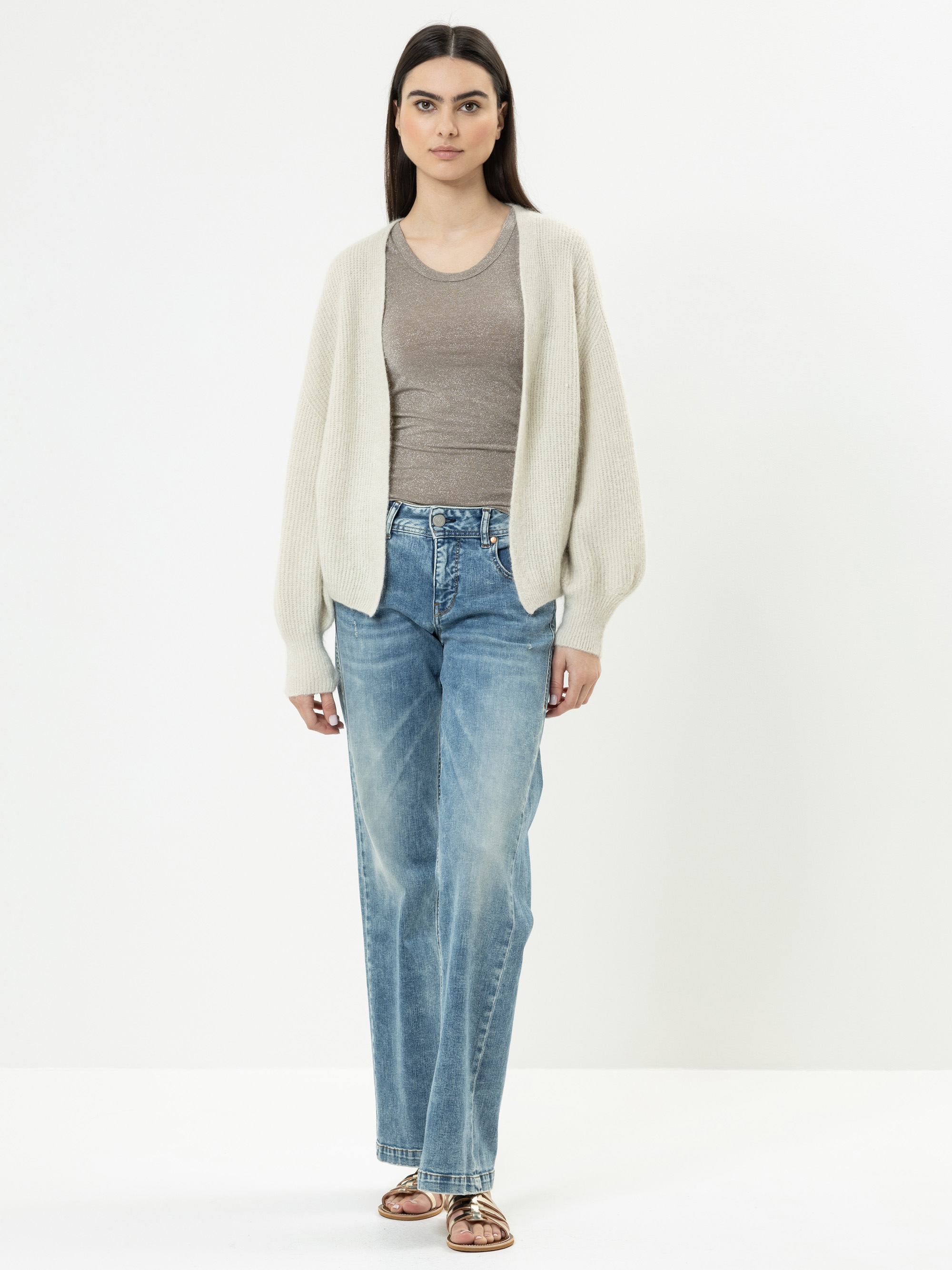 Eine Frau steht vor einem schlichten weißen Hintergrund und trägt eine hellbeige Strickjacke über einem taupefarbenen Oberteil, blaue Jeans und braune Sandalen. Sie hat langes dunkles Haar und einen neutralen Gesichtsausdruck.