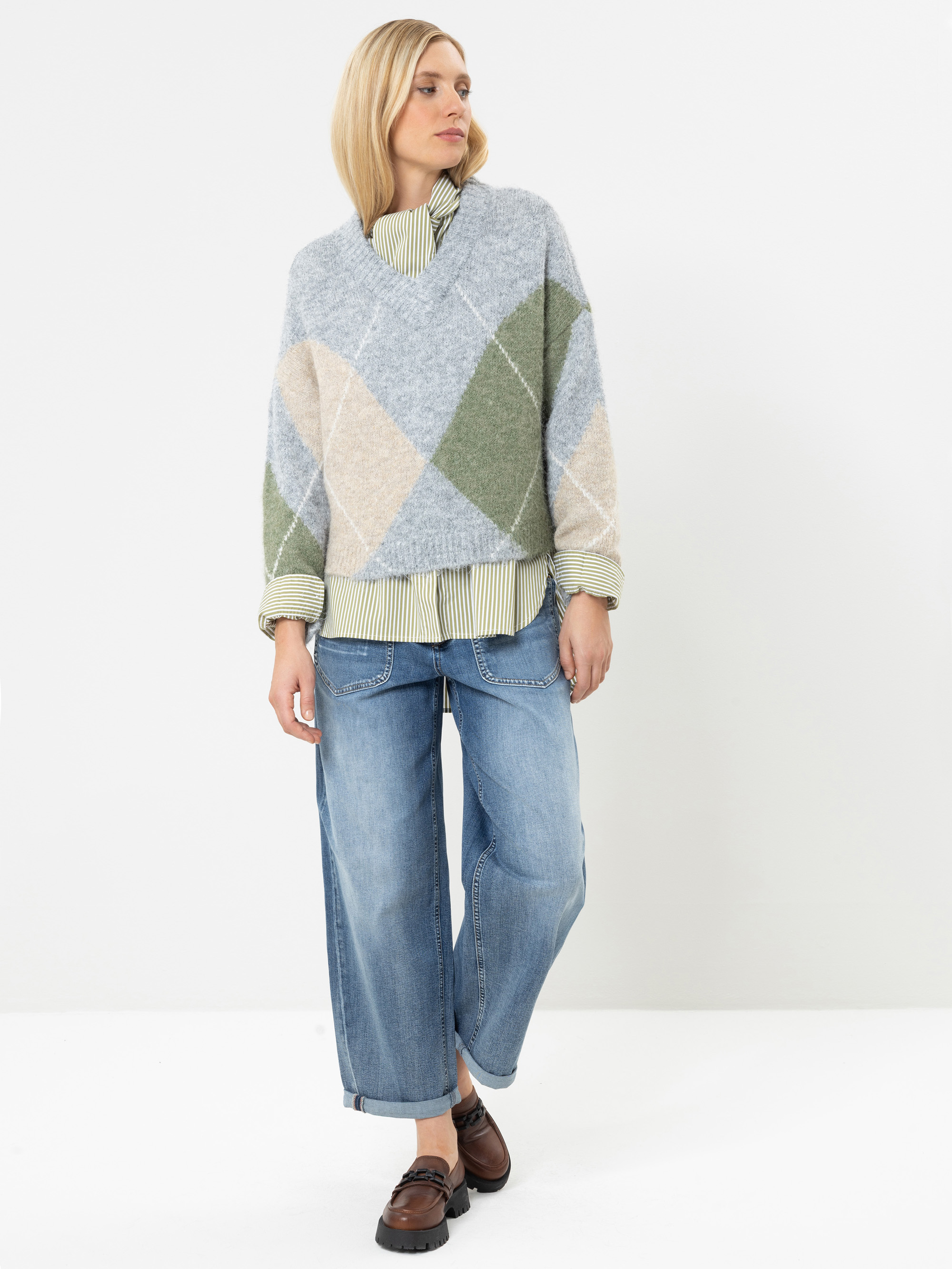 Eine Frau steht vor einem weißen Hintergrund und trägt einen hellblau-grünen Argyle-Pullover über einem gestreiften Hemd, eine lockere blaue Jeans und braune Slipper. Sie hat blondes Haar und blickt mit entspannter Körperhaltung zur Seite.
