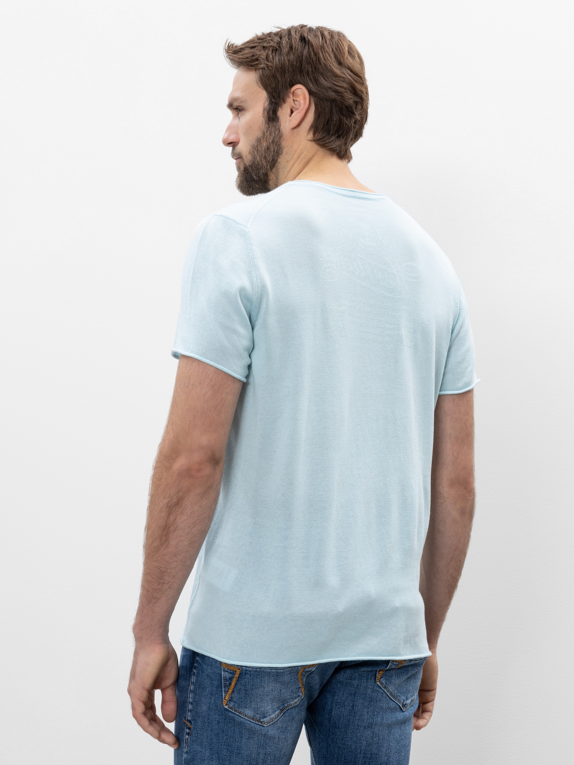 Ein Mann mit kurzen braunen Haaren und einem Bart steht und schaut von der Kamera weg. Er trägt ein hellblaues T-Shirt und eine blaue Jeans vor einem schlichten weißen Hintergrund.