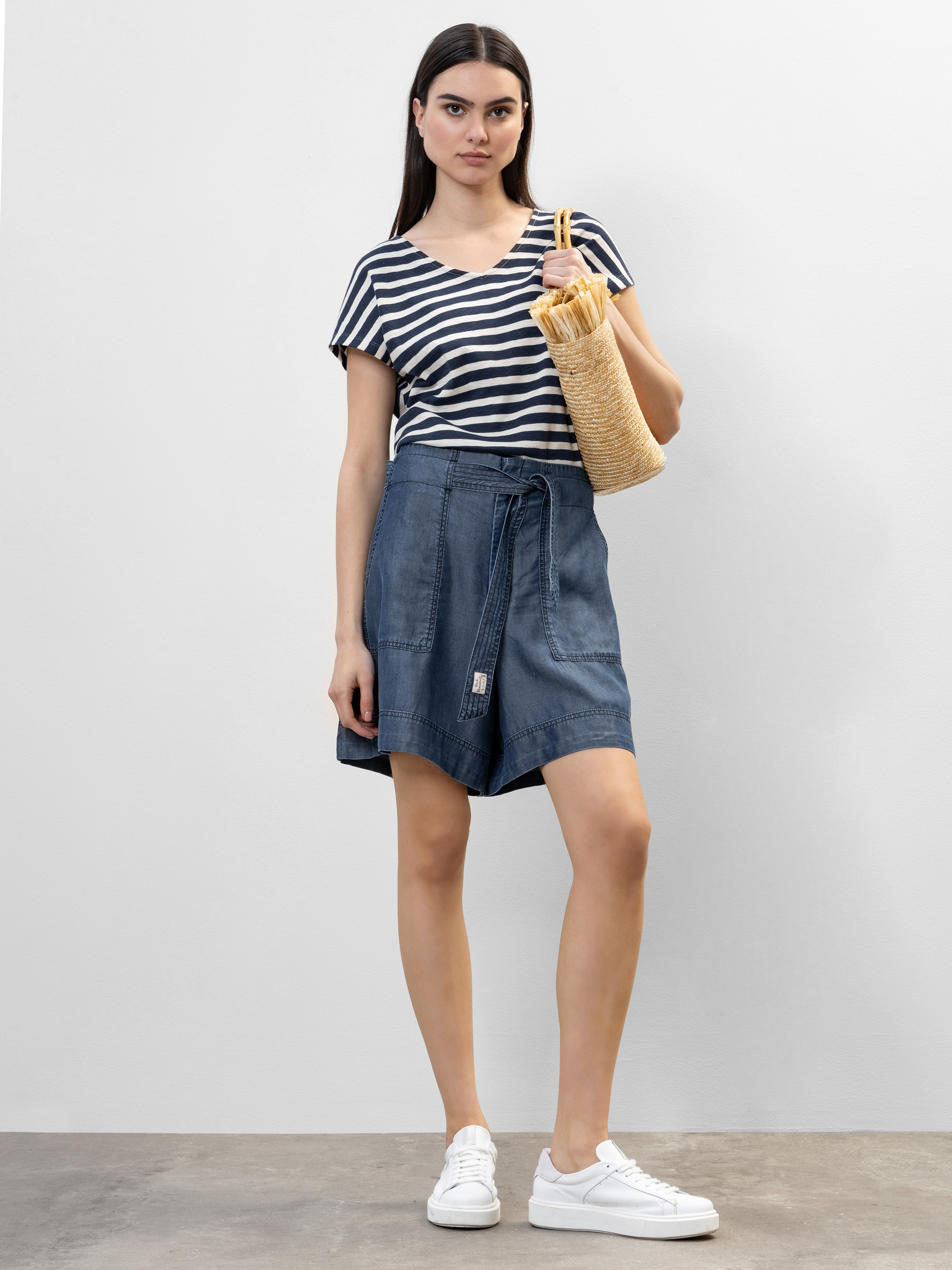 Eine Frau steht vor einem unifarbenen Hintergrund, trägt ein schwarz-weiß gestreiftes Oberteil, blaue Shorts mit hoher Taille, weiße Turnschuhe und hat eine geflochtene Strohtasche mit Holzgriffen dabei.