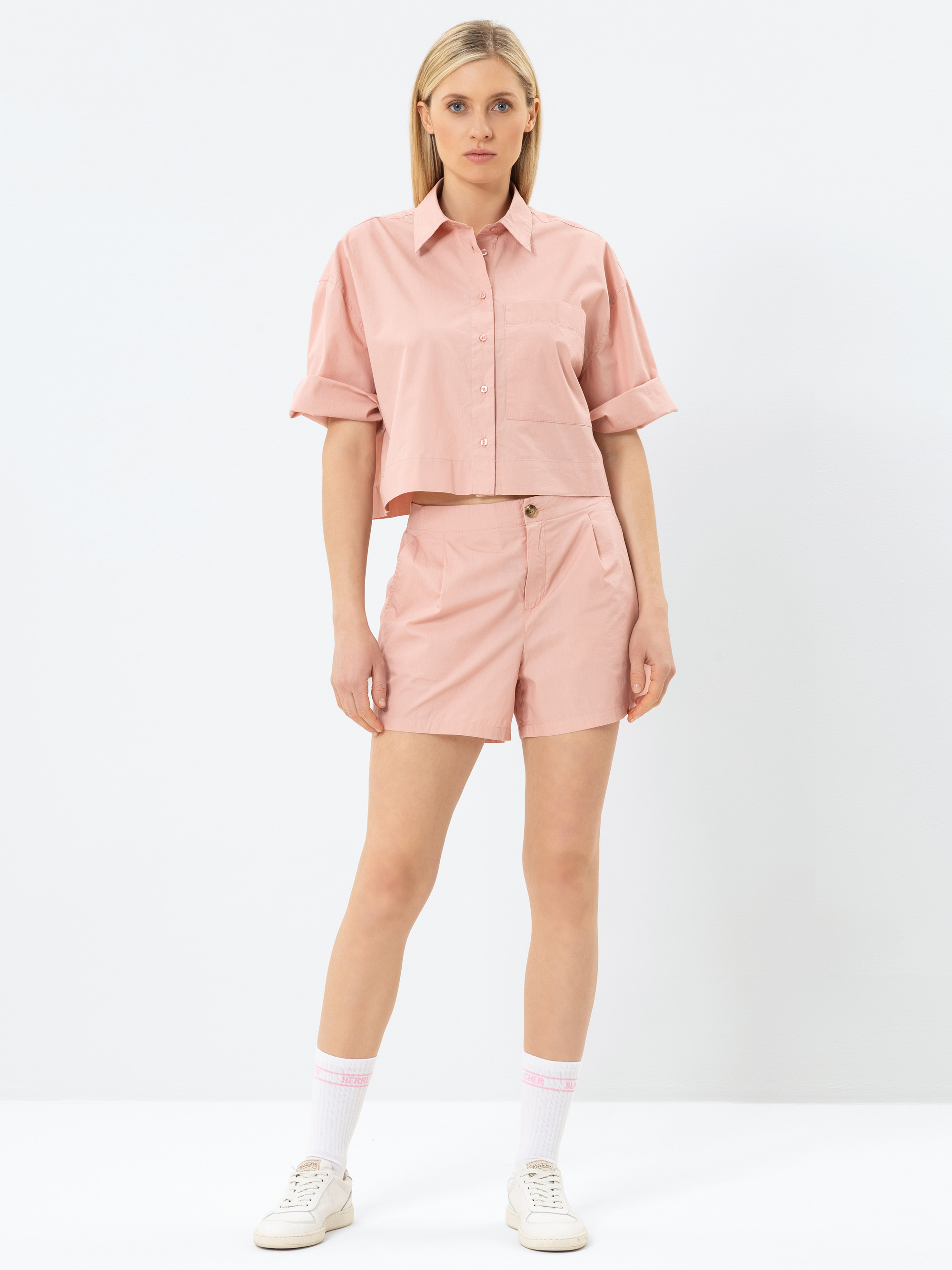 Eine Frau steht vor einem schlichten weißen Hintergrund und trägt ein hellrosa, kurzärmeliges Button-up-Hemd und passende Shorts, weiße Socken mit rosa Streifen und weiße Turnschuhe. Sie hat glattes blondes Haar und einen neutralen Gesichtsausdruck.