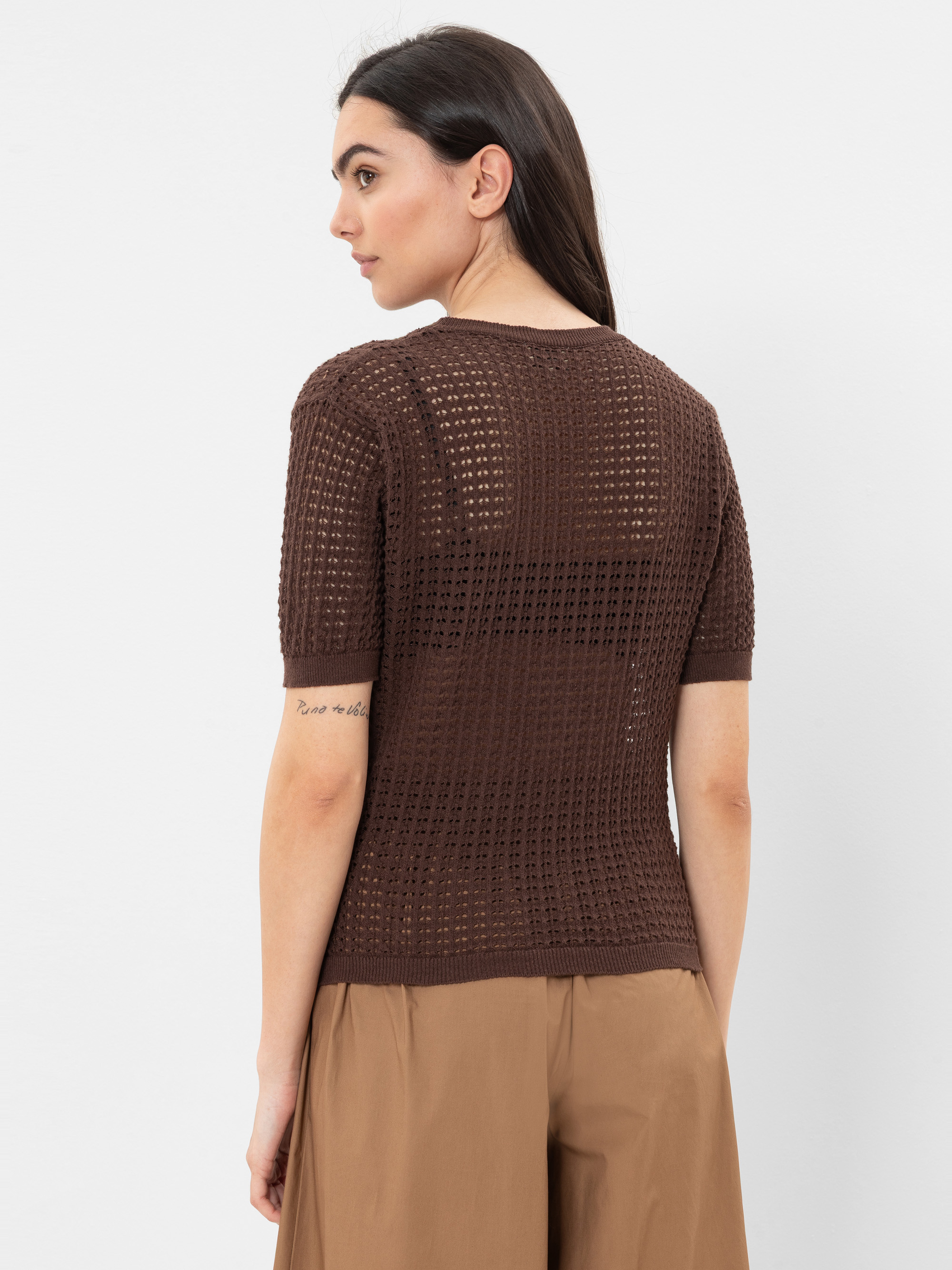 Eine Frau mit langen dunklen Haaren, bekleidet mit einem braunen kurzärmeligen Strickoberteil und einer hellbraunen Hose, steht schräg gegenüber und zeigt eine kursive Tätowierung auf ihrem rechten Unterarm vor einem schlichten weißen Hintergrund.