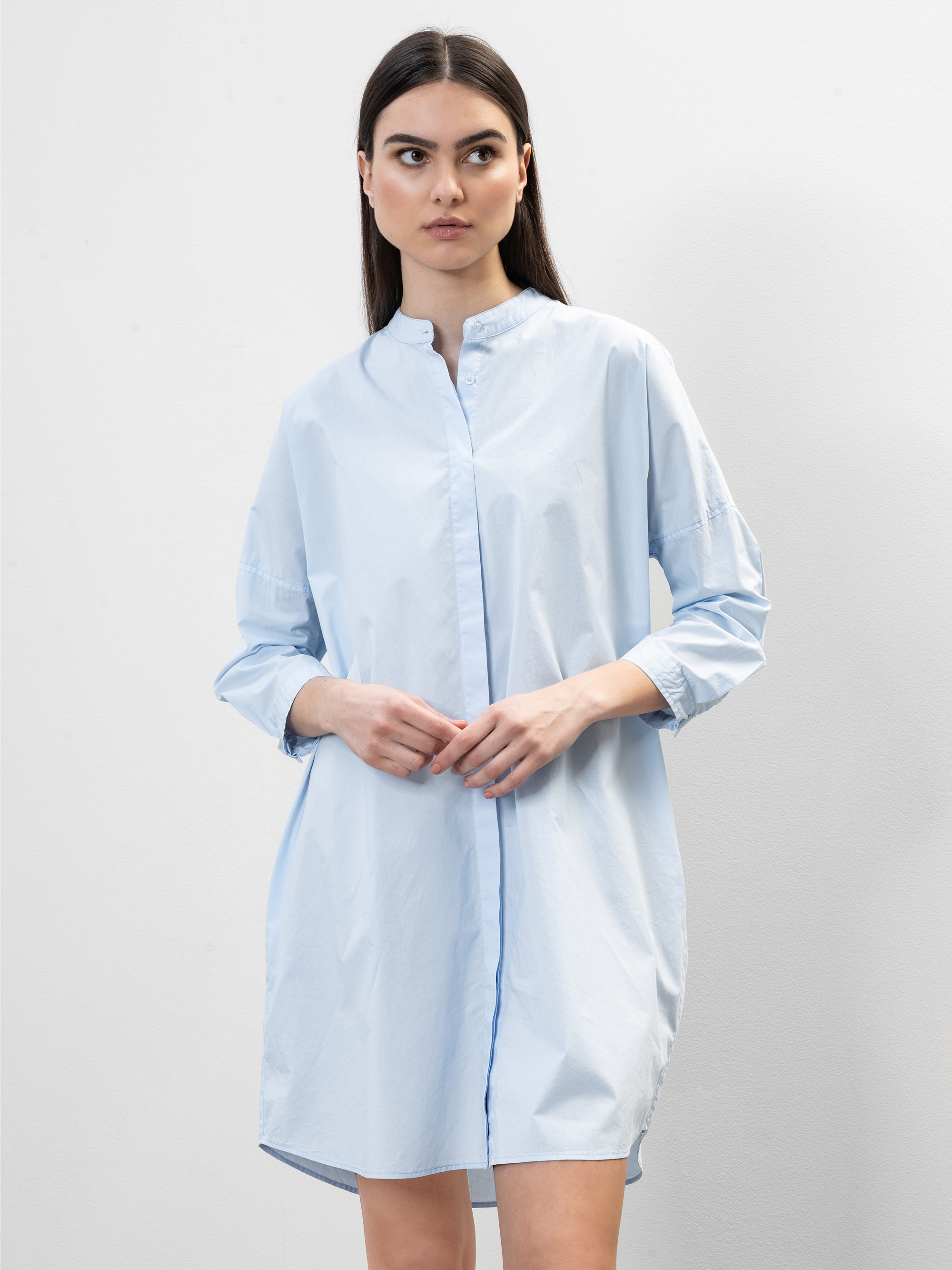 Eine Frau mit langen dunklen Haaren, die ein lockeres hellblaues Hemdkleid mit Dreiviertelärmeln trägt, steht vor einem schlichten hellen Hintergrund und blickt mit neutraler Miene leicht zur Seite.