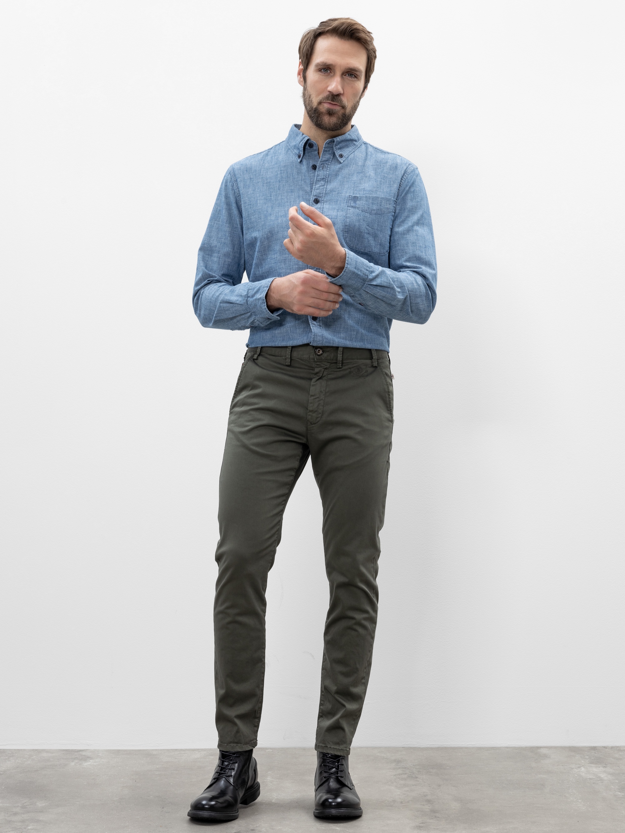 Ein Mann steht vor einem schlichten weißen Hintergrund und trägt ein hellblaues Button-up-Hemd, eine olivgrüne Hose und schwarze Schuhe. Er rückt seine linke Hemdmanschette zurecht und schaut geradeaus.