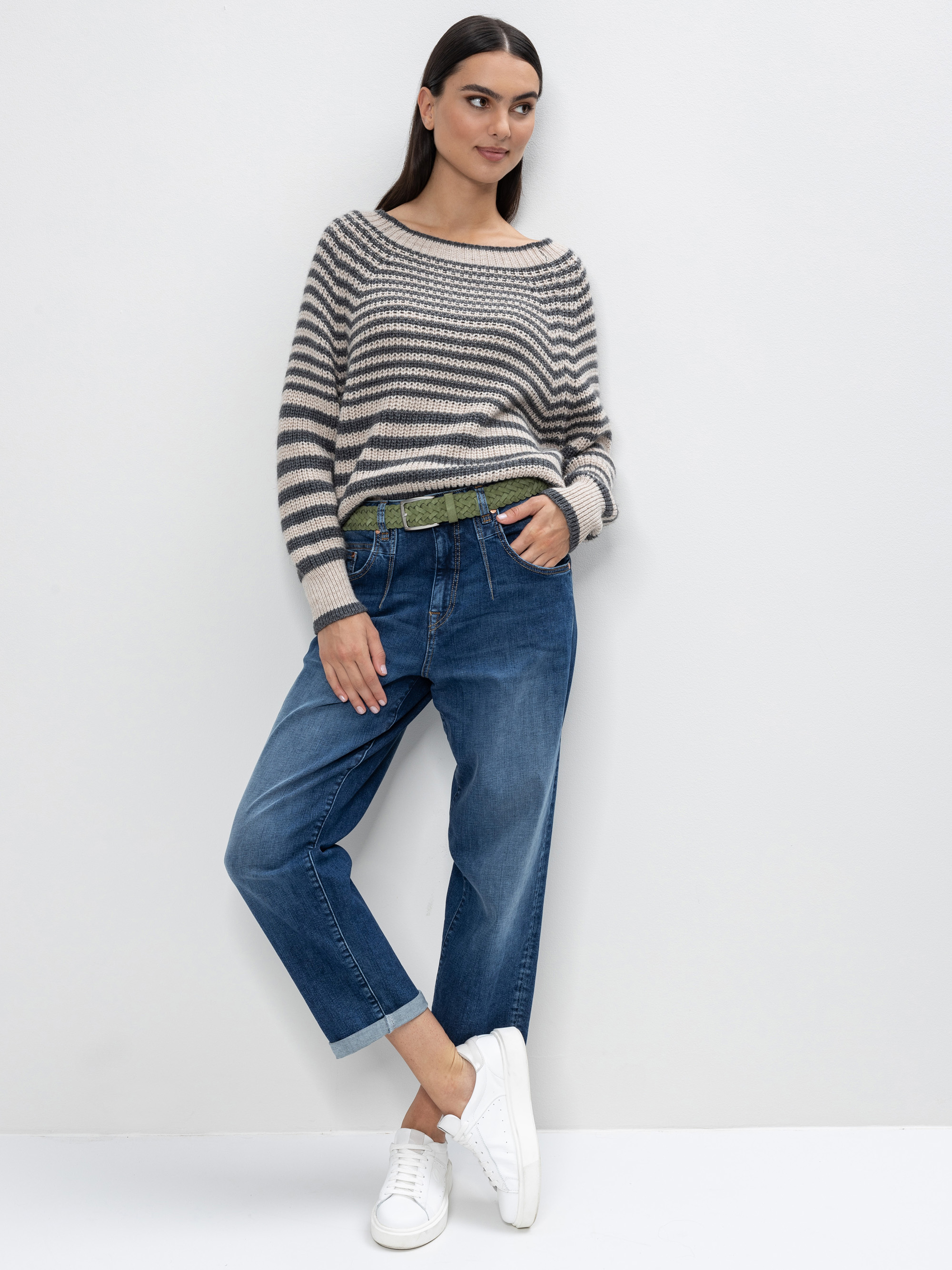 Eine Frau steht vor einer weißen Wand, trägt einen gestreiften Pullover, lässige Bluejeans mit aufgerollten Manschetten, einen grünen Gürtel und weiße Turnschuhe, eine Hand in der Tasche und die Haare offen.