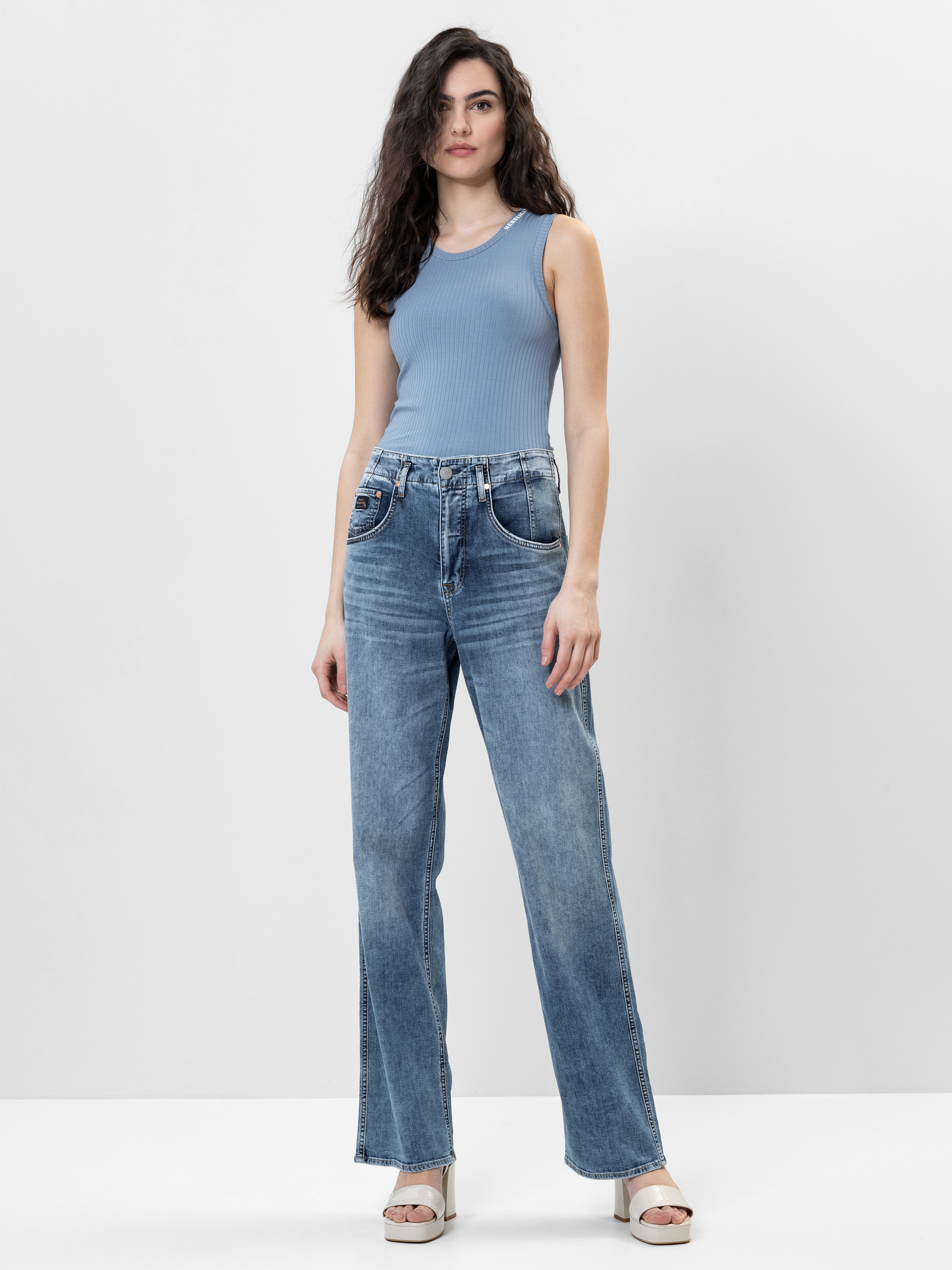 Eine Frau mit langen, gewellten Haaren trägt ein hellblaues, ärmelloses, geripptes Oberteil, hochtaillierte Blue Jeans und weiße Plateausandalen vor einem schlichten weißen Hintergrund.